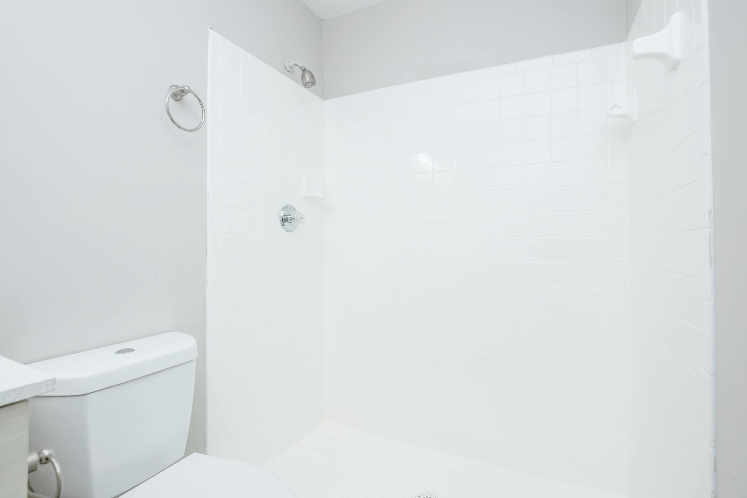 A clean, modern bathroom featuring a white tiled shower area with a chrome showerhead, a towel ring on the wall, and a white toilet in the corner. The walls are painted light gray, creating a bright and spacious appearance.