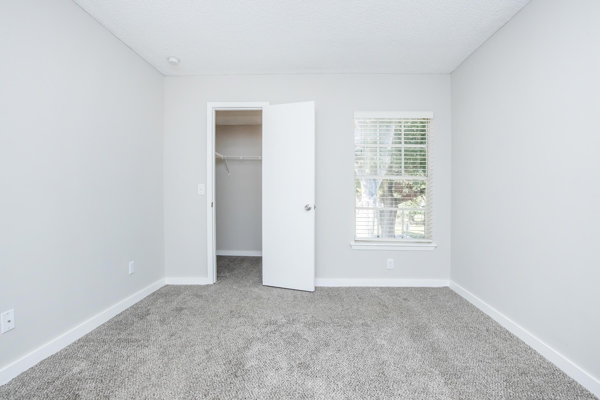 A clean, empty room with light gray walls and carpet. There is a closed closet with an open door on one side and a window with blinds allowing natural light. The overall space appears well-maintained and bright, suitable for various uses or furnishings.