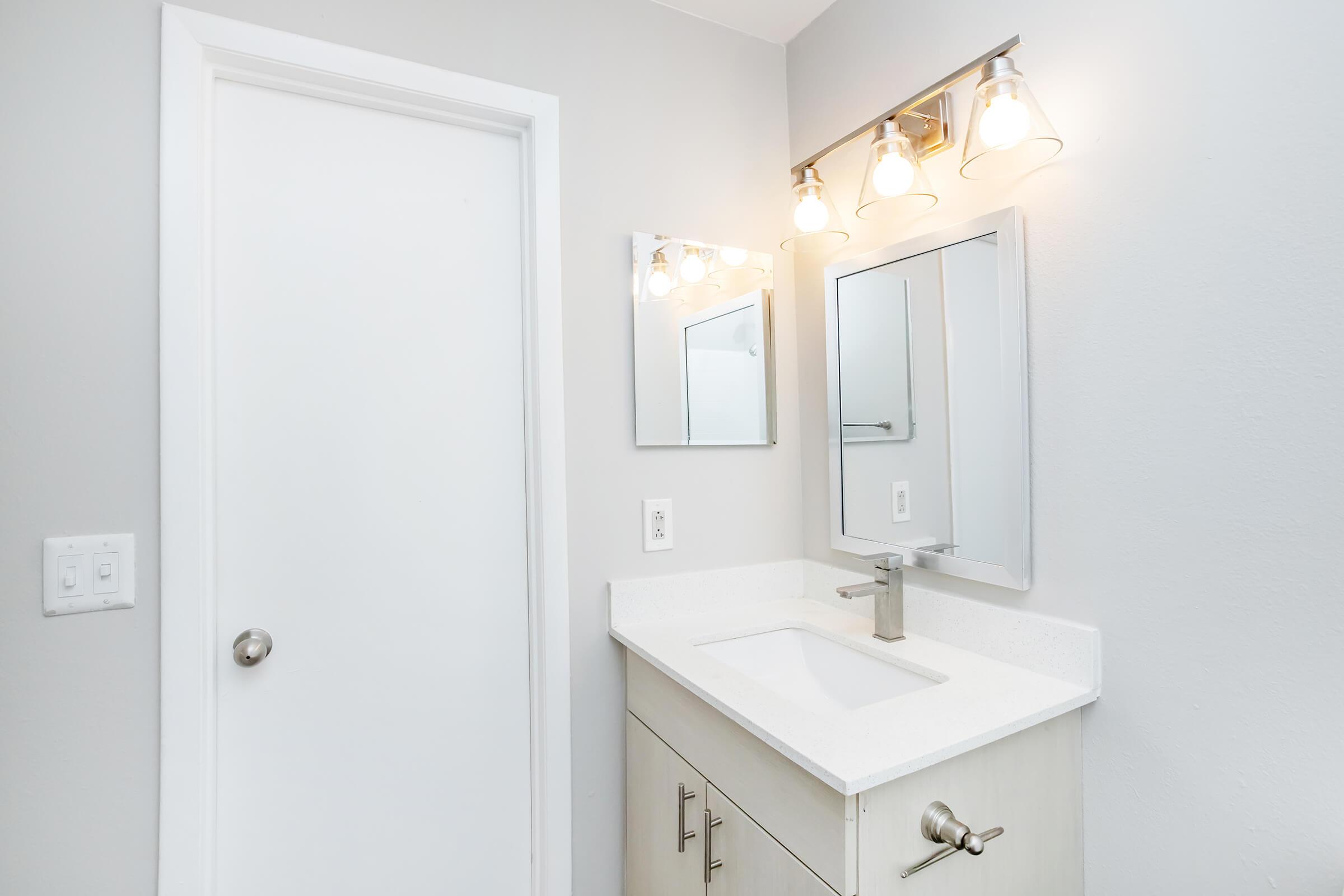 Modern bathroom featuring a white sink with a sleek faucet, light-colored cabinetry, a large mirror, and a light fixture with three bulbs. The walls are painted in a soft gray, and a closed door is visible on the left side. Natural light enhances the clean, minimalist aesthetic.