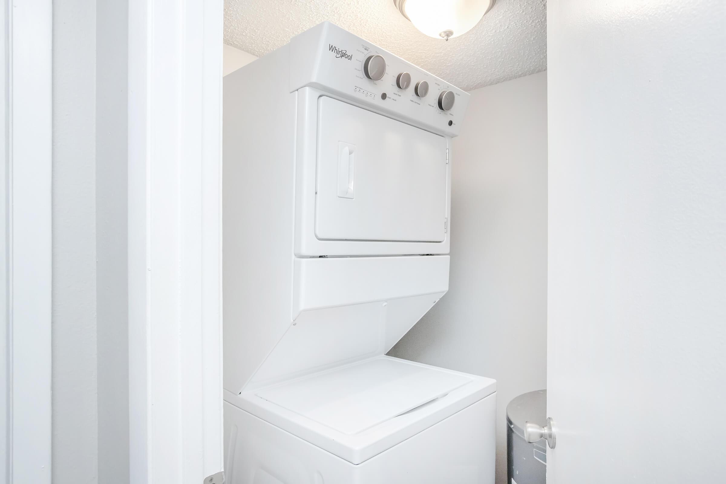 A stacked white washer and dryer unit in a small laundry space, with a light fixture above. The walls are painted a light color, and there is a gray trash can in the corner. The door is slightly ajar, providing a view of the appliances.