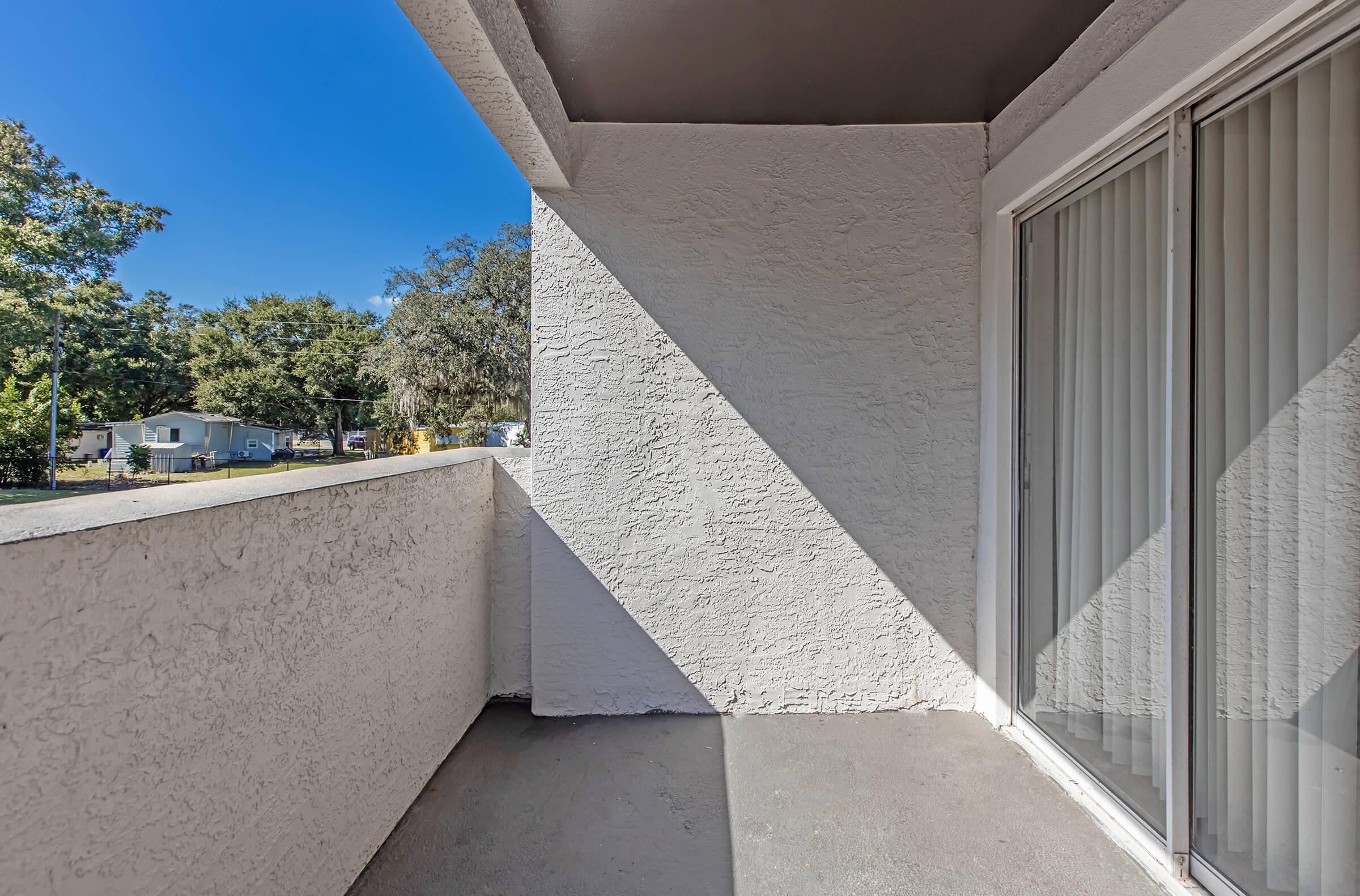 View of a vacant balcony with a textured white wall and sliding glass door. Sunlight casts sharp shadows on the concrete floor, while trees and houses are visible in the background. The clear blue sky suggests a bright, sunny day.