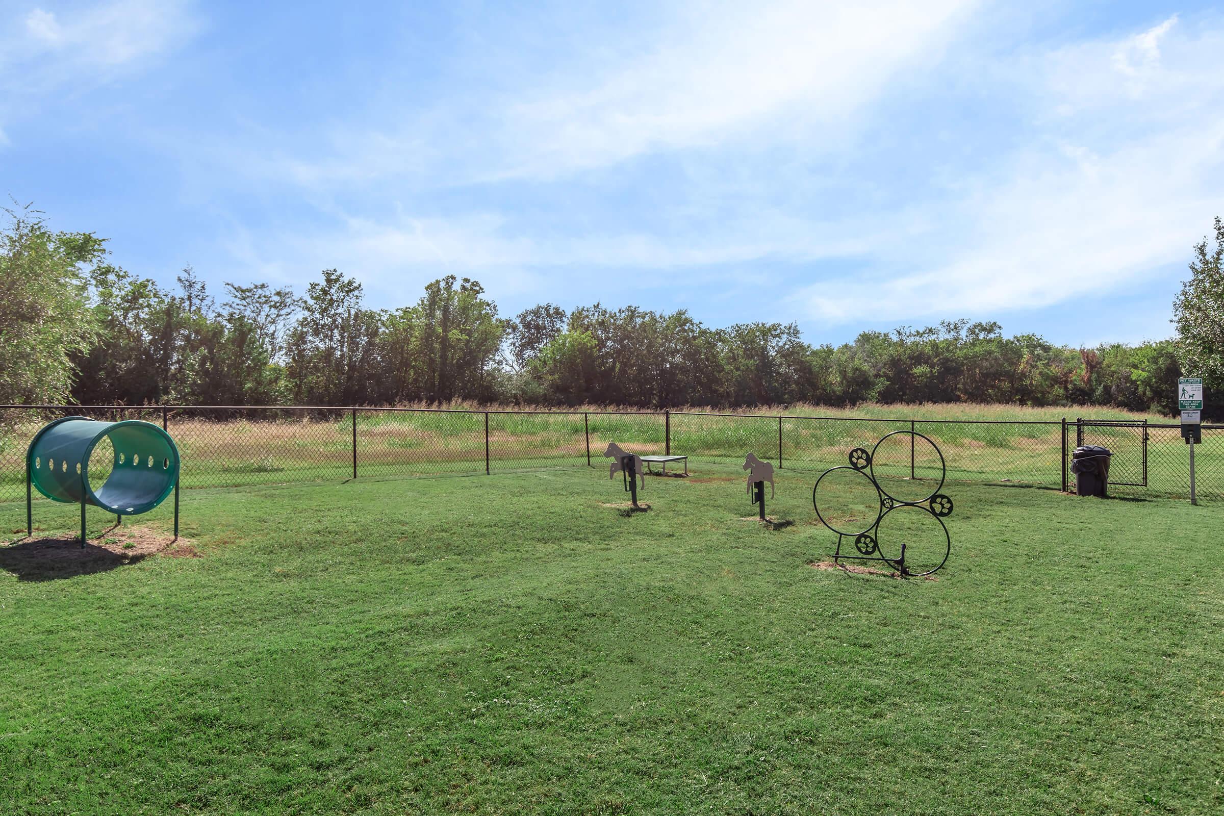A grassy dog park featuring a green tunnel for pets to play in, a water station, and a circular obstacle course structure. The area is enclosed with a black chain-link fence, surrounded by trees and open fields in the background under a clear blue sky.