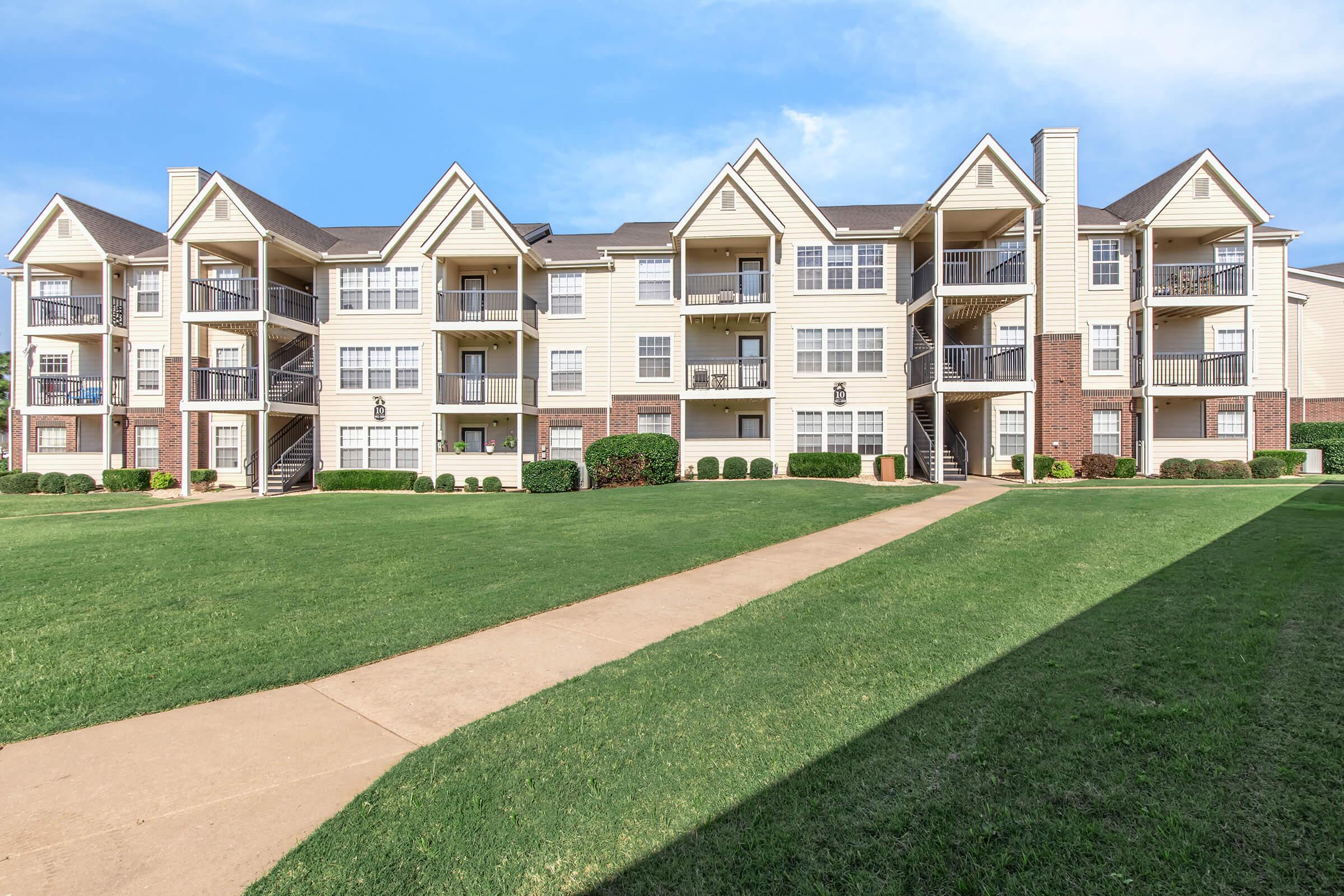 A three-story apartment building with a light-colored exterior and multiple balconies, surrounded by well-maintained green lawns and pathways. The building features a symmetrical design with peaks on the roof and is set against a clear blue sky.