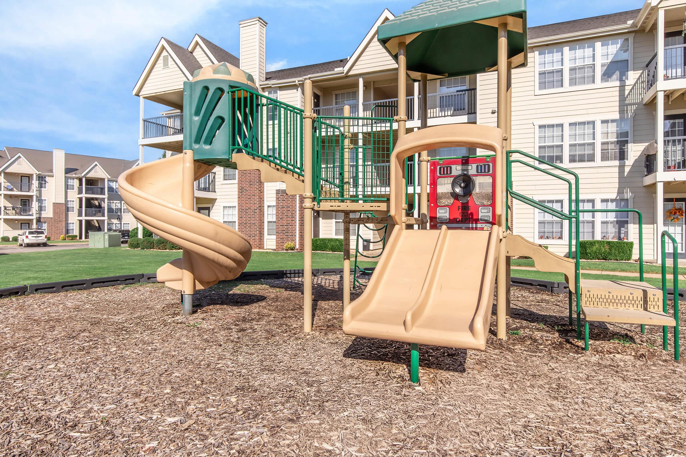 Playground equipment featuring a beige slide, a spiral slide, and climbing structures, set on mulch. In the background, a residential building with balconies is visible under a blue sky. The playground is designed for children’s outdoor play.