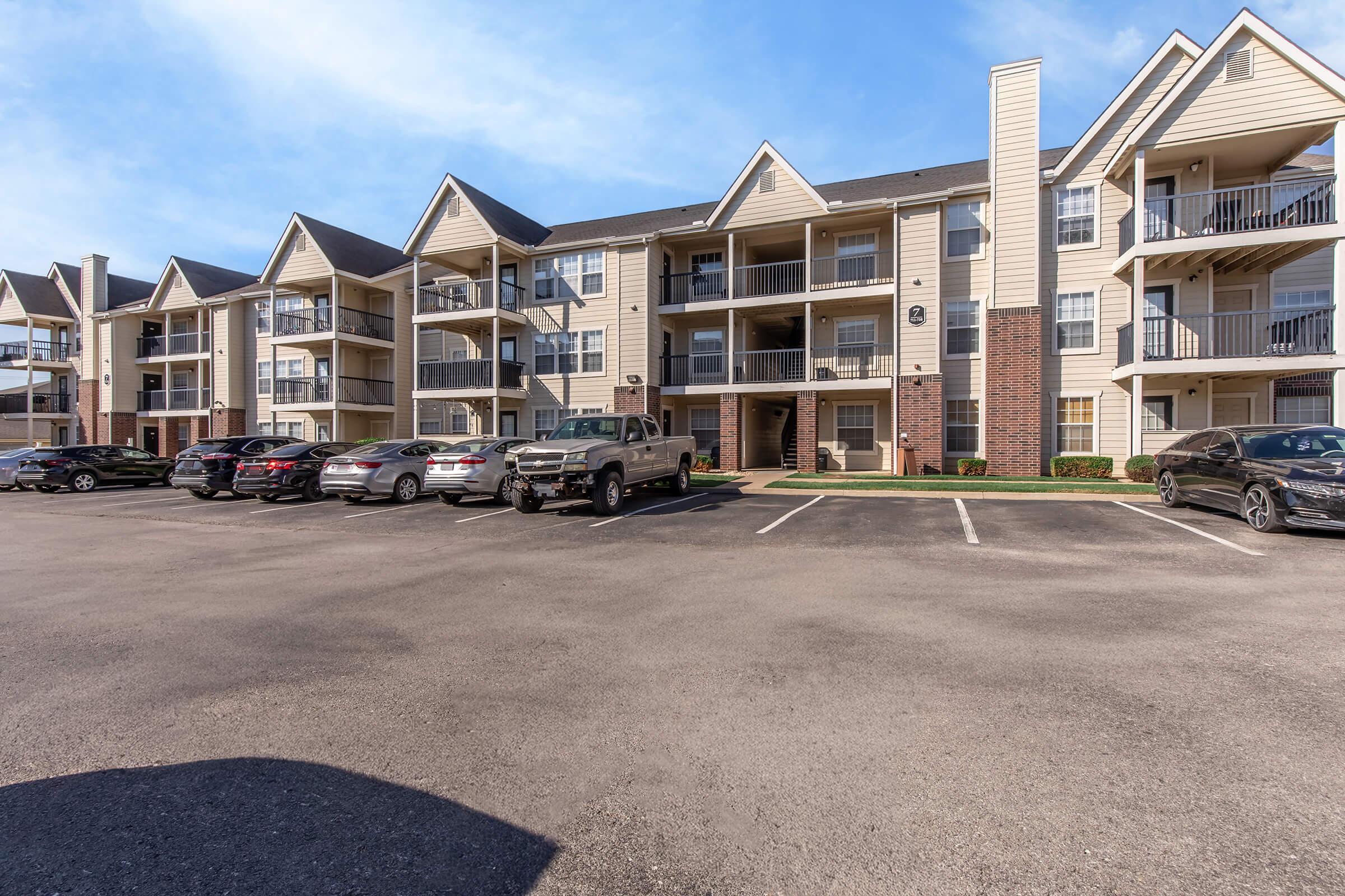 A multi-story apartment complex with a modern design, featuring balconies and a parking lot with several vehicles. The building is light-colored with dark roofs, and there is clear blue sky in the background. The setting appears well-maintained and inviting.
