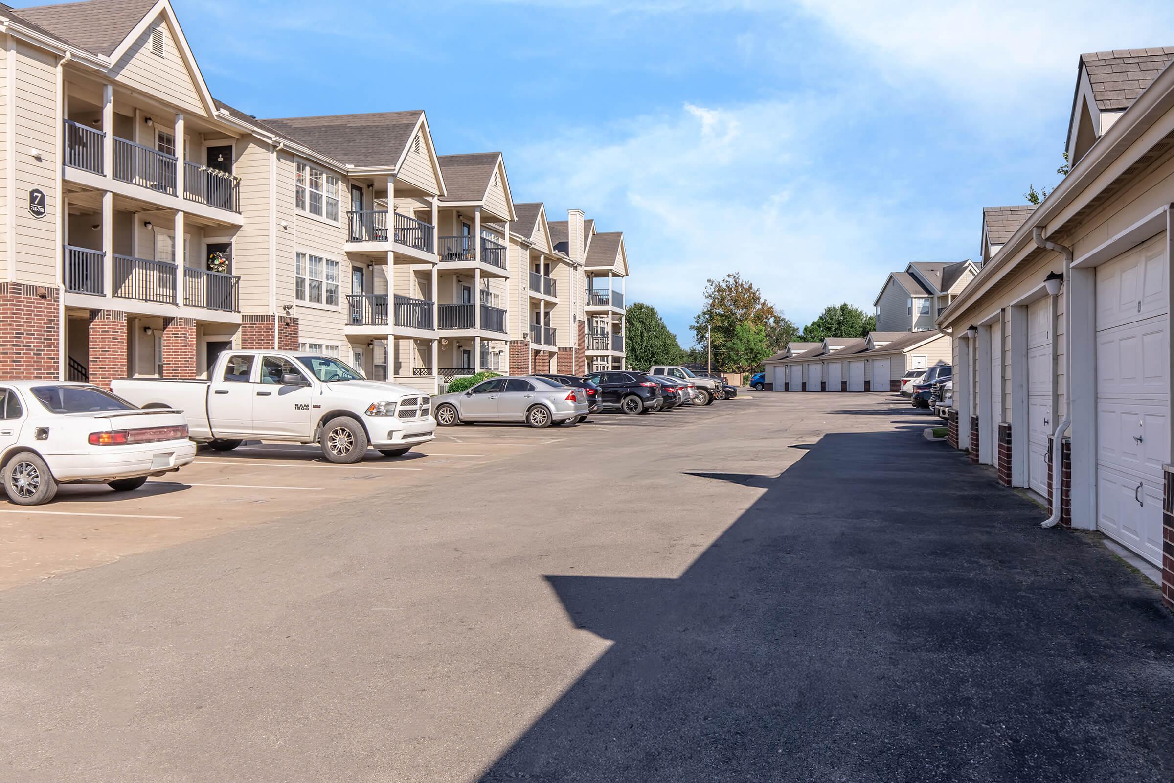 A view of a multi-unit residential complex featuring several three-story buildings with balconies. A paved parking lot is visible with various cars parked along the side. On the right, there are garage units, and the scene is set against a clear blue sky and greenery in the background.