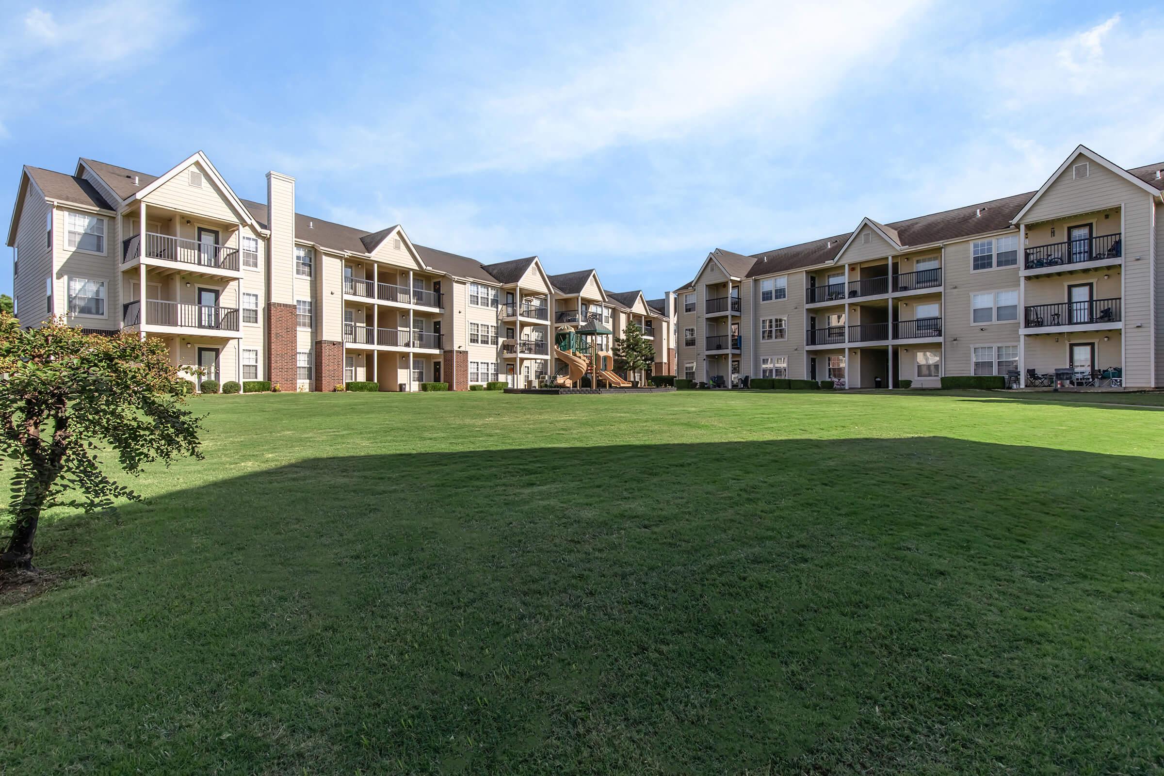 A scenic view of two multi-unit apartment buildings surrounded by a spacious green lawn. The buildings feature balconies and patios, with various trees and landscaping elements in the foreground. The sky is clear, indicating a bright and sunny day.
