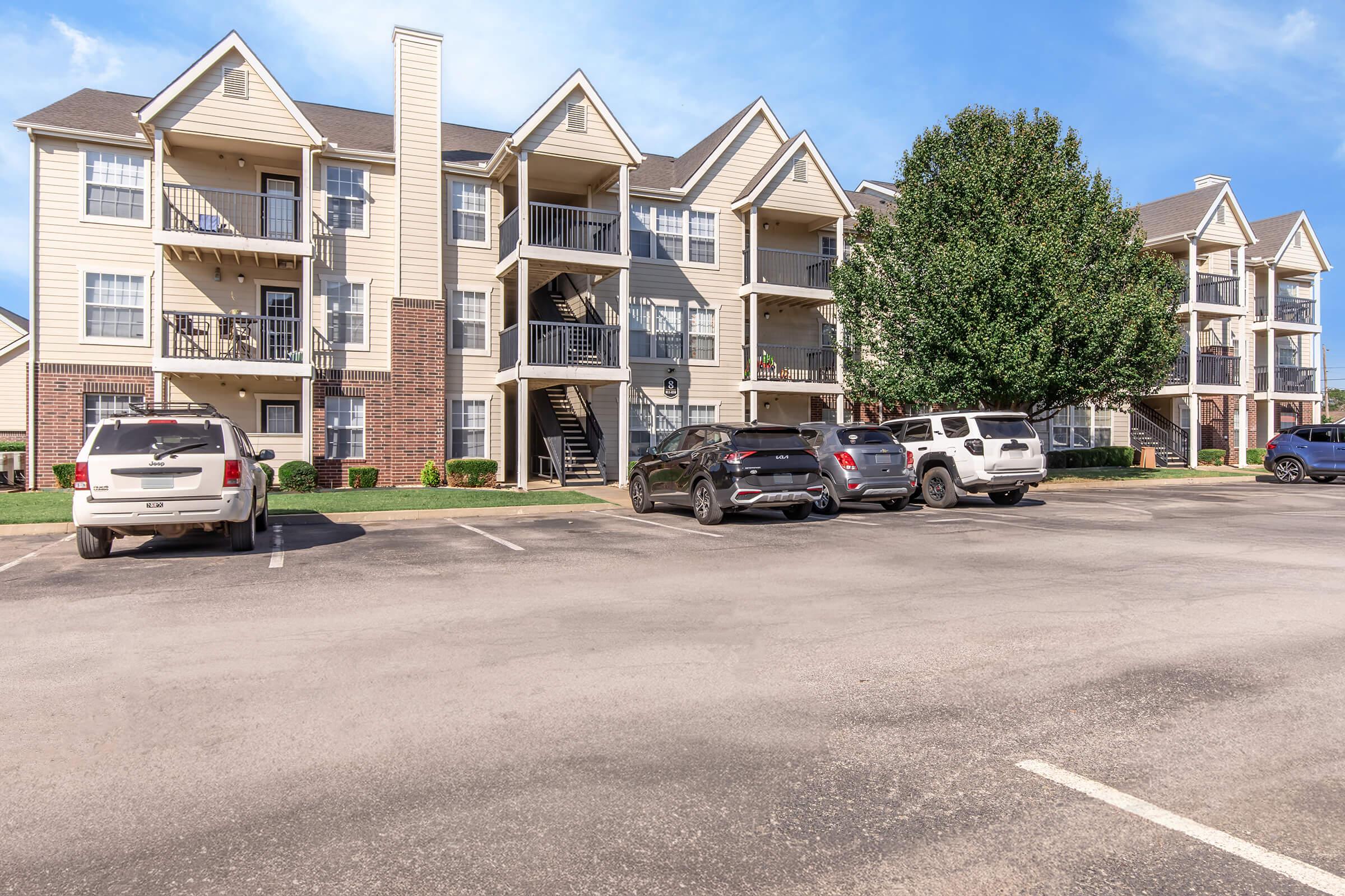 A spacious residential apartment complex featuring two buildings with multiple balconies, surrounded by green grass. Several parked cars are visible in the foreground, and a large tree is present next to the buildings under a clear blue sky.