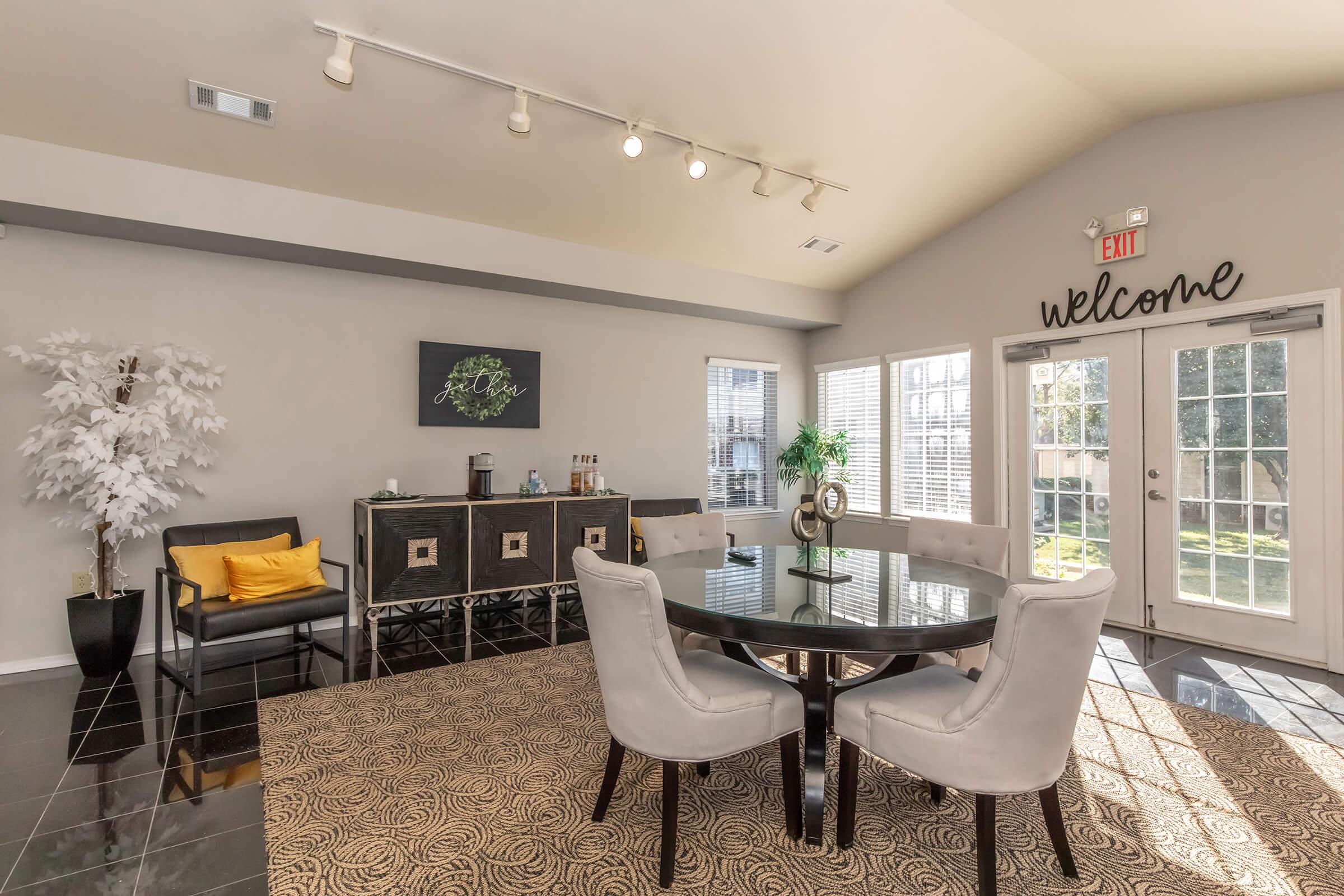 A modern, stylish lounge area featuring a round table surrounded by gray chairs, a black sideboard with decorative items, and a faux white tree. Large windows allow natural light in, with a "welcome" sign on the wall. The floor has an intricate patterned rug, creating an inviting atmosphere.