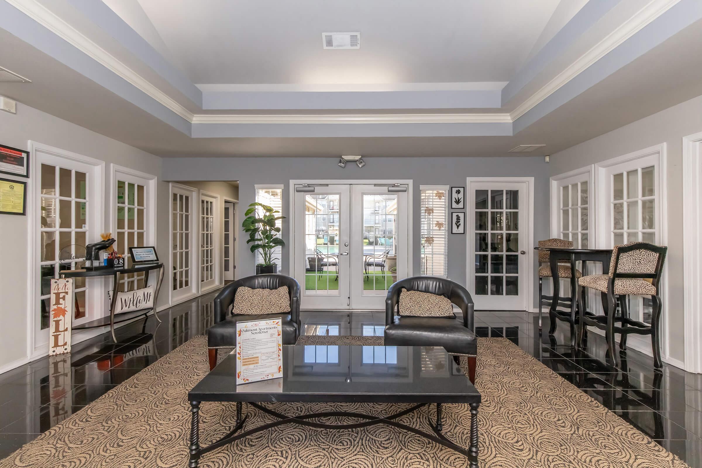 A modern lobby area featuring black tiled flooring, seating with patterned cushions, and a coffee table. Large windows and double doors provide natural light, while a potted plant adds a touch of greenery. Informational displays and bar-height seating are visible in the background.