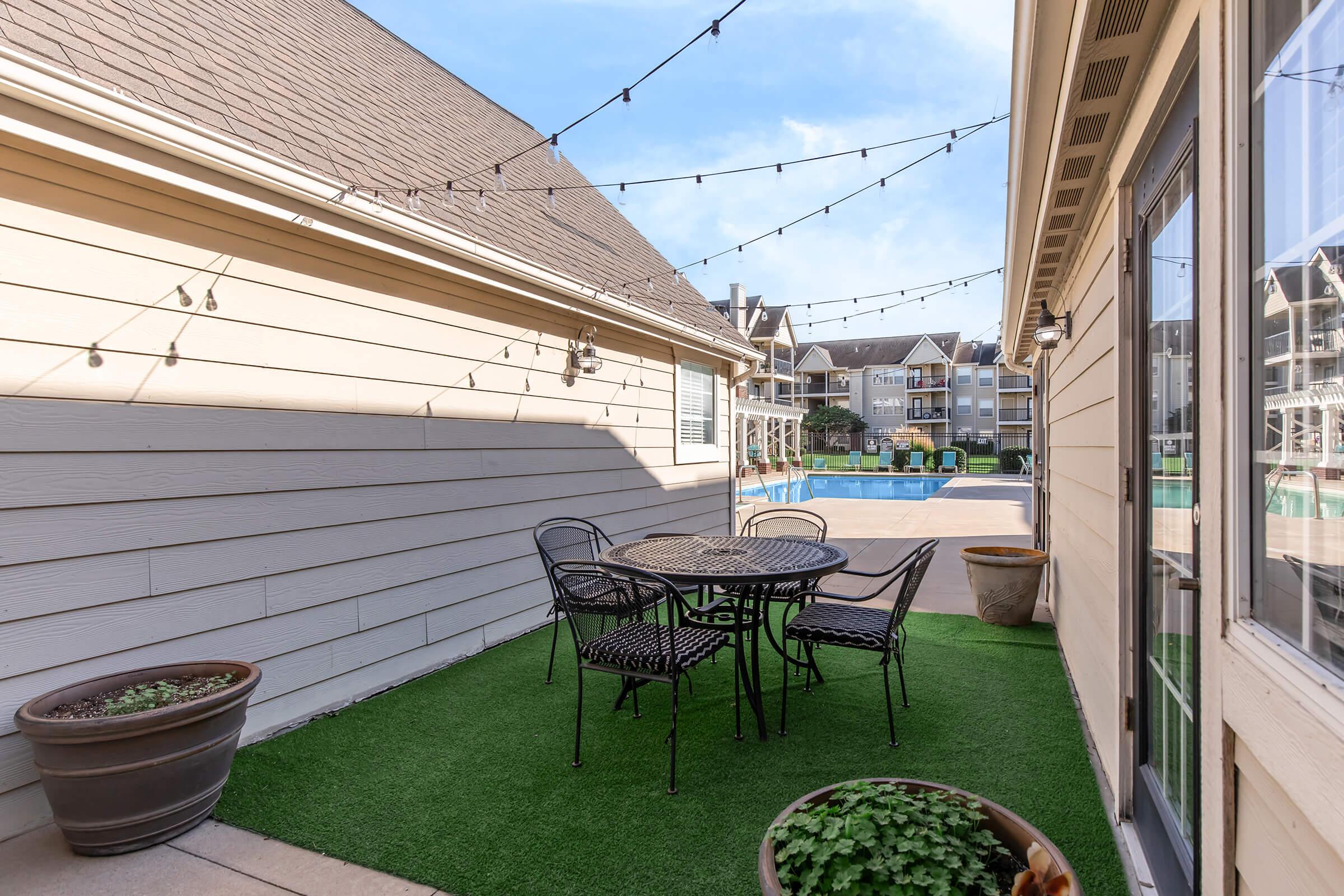 A small outdoor patio with a round table and four black wrought iron chairs, surrounded by planters. The space features artificial grass and is lined with string lights above. In the background, a swimming pool is visible, along with a view of neighboring apartment buildings.