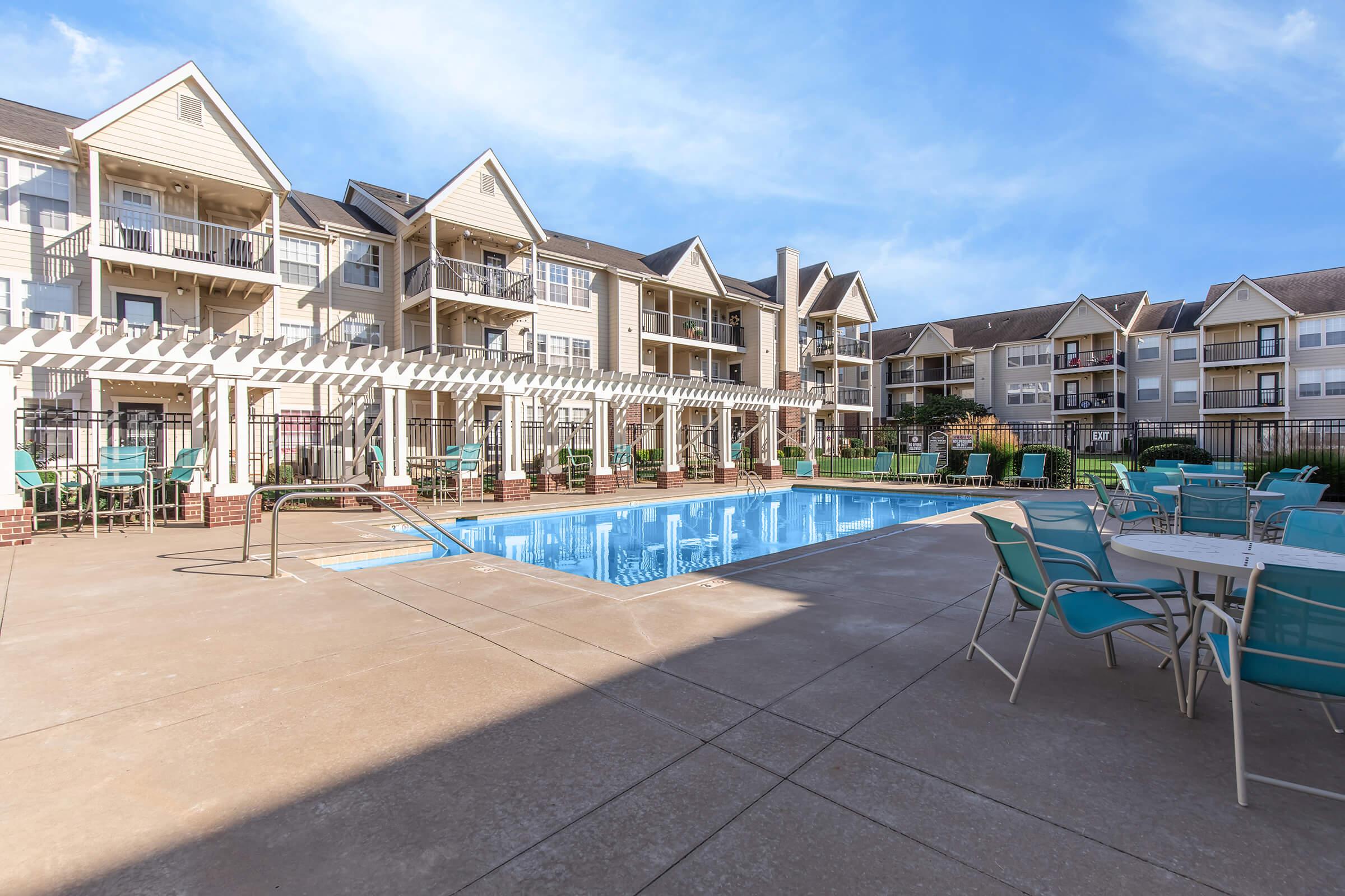 Swimming pool area surrounded by multi-story apartment buildings. The pool is flanked by lounge chairs and tables under a pergola, with clear blue skies overhead, creating a relaxing outdoor space.