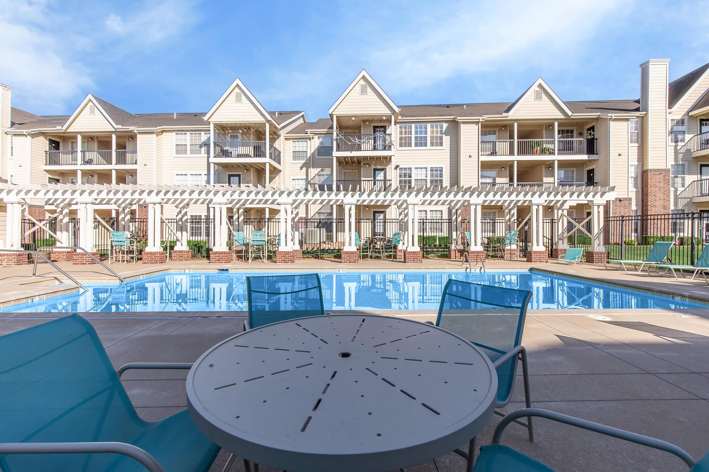 View of an outdoor swimming pool surrounded by lounge chairs and an outdoor dining table. In the background, a multi-story apartment building with balconies is visible, along with a blue sky and scattered clouds. The scene is bright and inviting, ideal for relaxation.