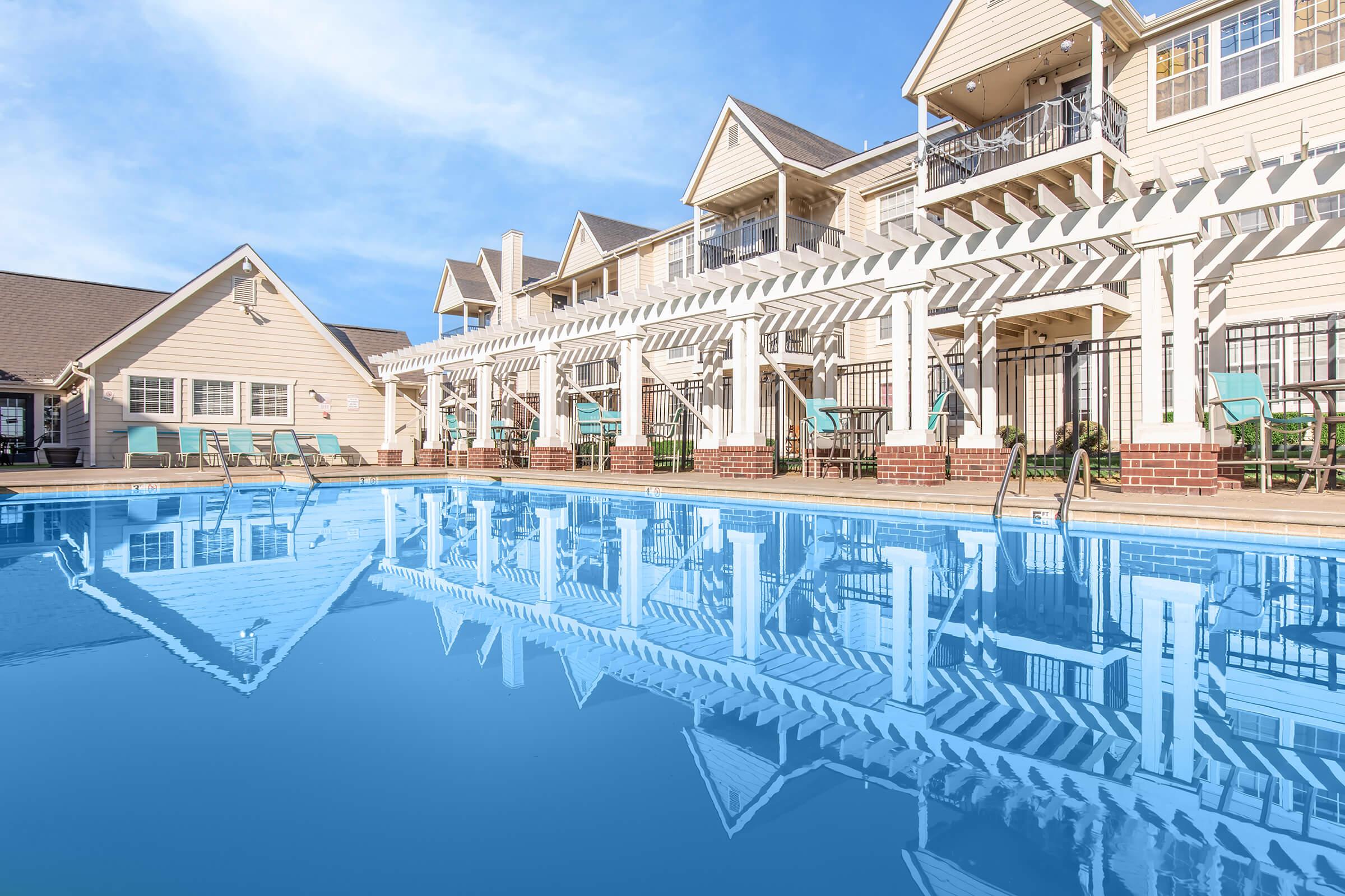 A tranquil pool area reflecting a picturesque building with multiple stories. The pool is surrounded by lounge chairs, and there are shaded seating spaces underneath a pergola. Clear blue sky overhead enhances the serene atmosphere of this inviting outdoor setting.