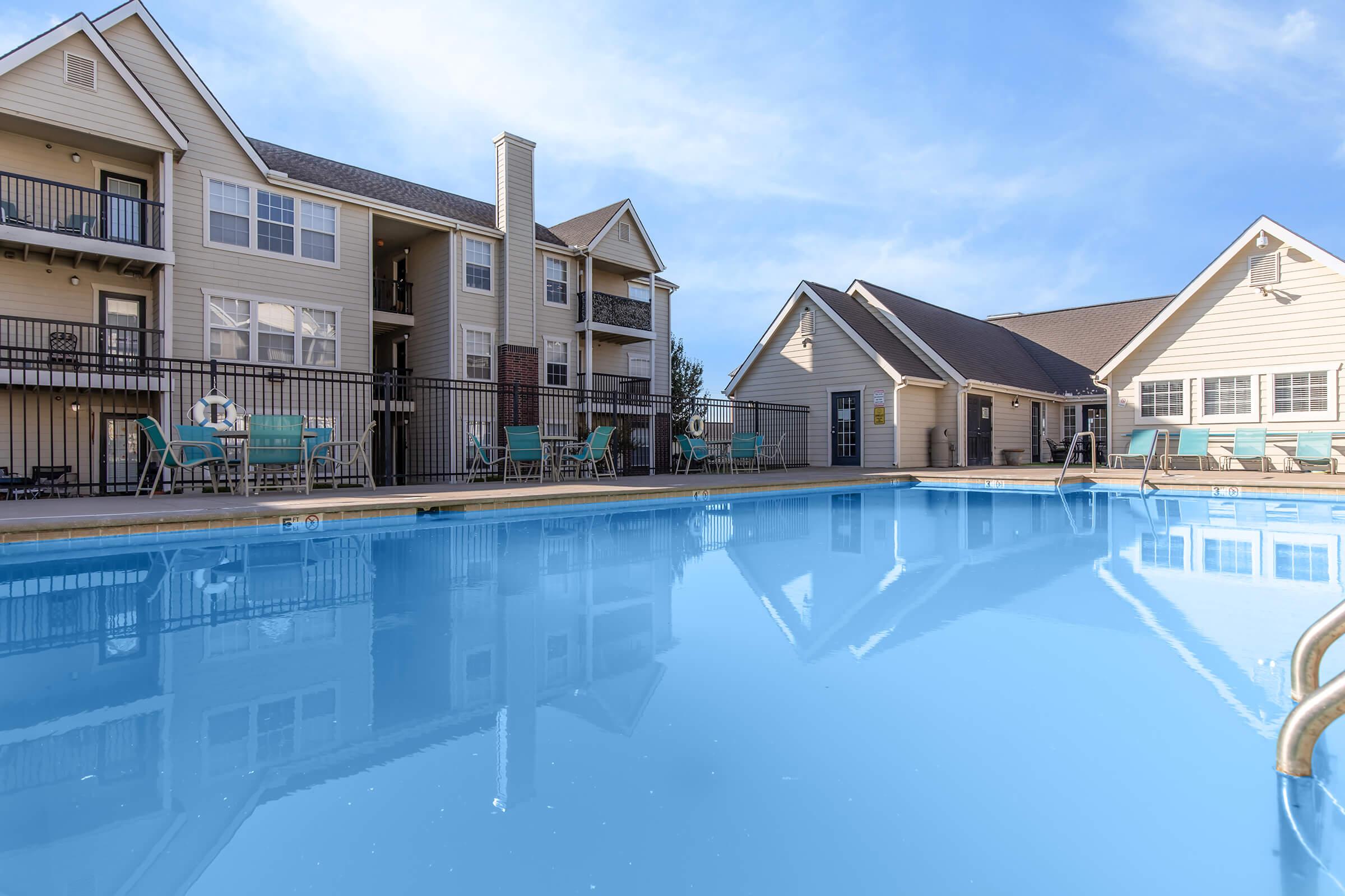 A clear blue swimming pool reflecting nearby residential buildings and a clubhouse under a bright sky. The pool area features lounge chairs and tables, with the structure of the apartments visible in the background. The scene conveys a calm and inviting atmosphere.