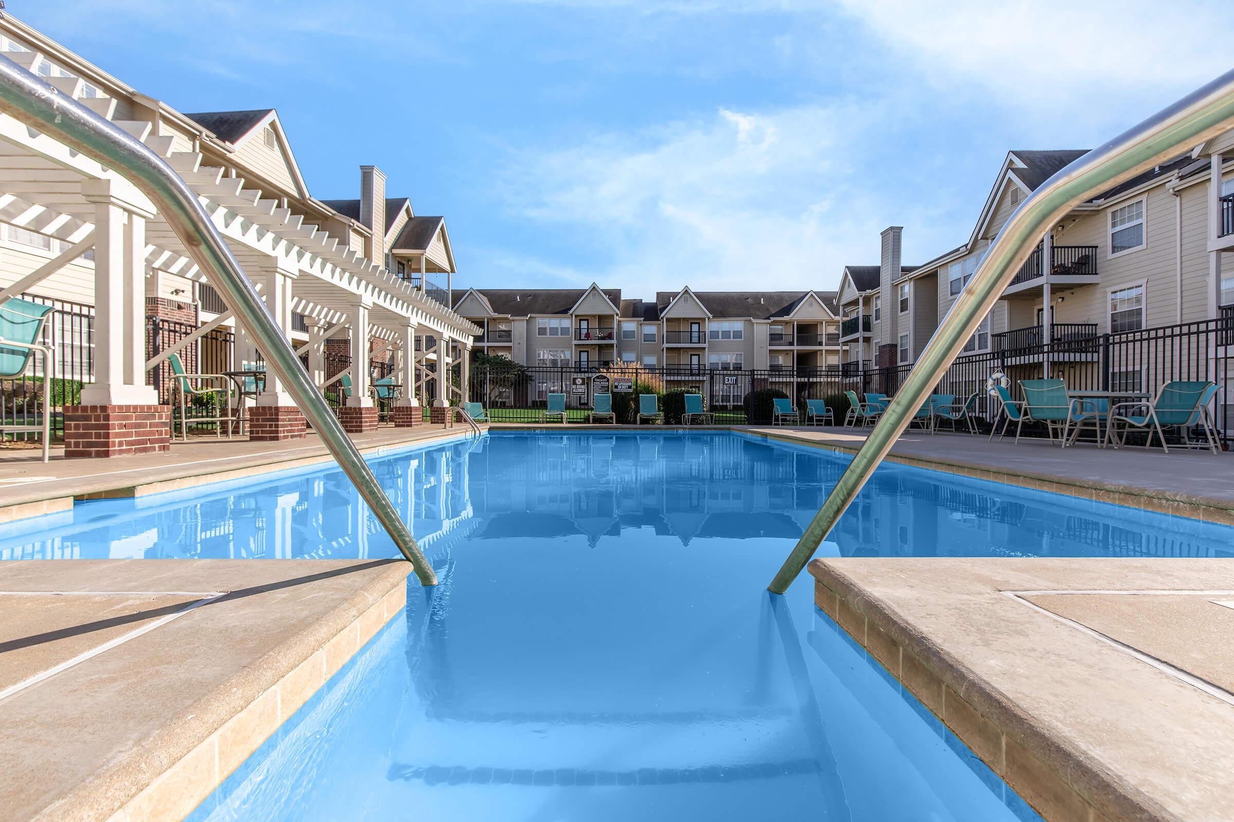 A clear blue swimming pool with a shallow entrance, framed by metal railings. In the background, there are several residential apartment buildings and a pergola with green chairs scattered around the pool area. The sky is bright with a few clouds.