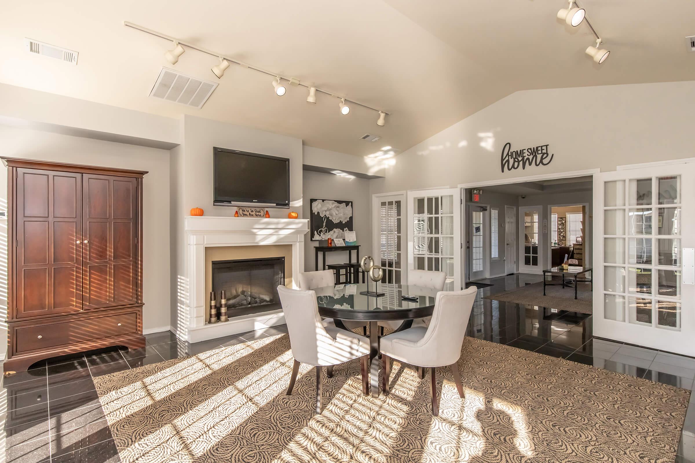 A modern living space featuring a round black dining table with two upholstered chairs, a stylish cabinet, and a wall-mounted TV above a fireplace. Large windows allow natural light to illuminate the room, casting shadows on the patterned rug. The decor includes seasonal accents, creating a warm, inviting atmosphere.