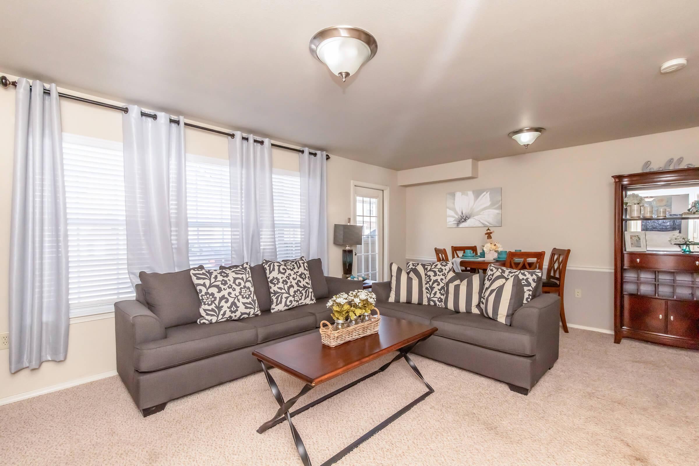 A cozy living room featuring two gray sofas adorned with patterned pillows, a wooden coffee table in the center, and light gray curtains framing the windows. A small dining area with a round table and chairs is visible in the background, along with a decorative cabinet. The space is well-lit and inviting.