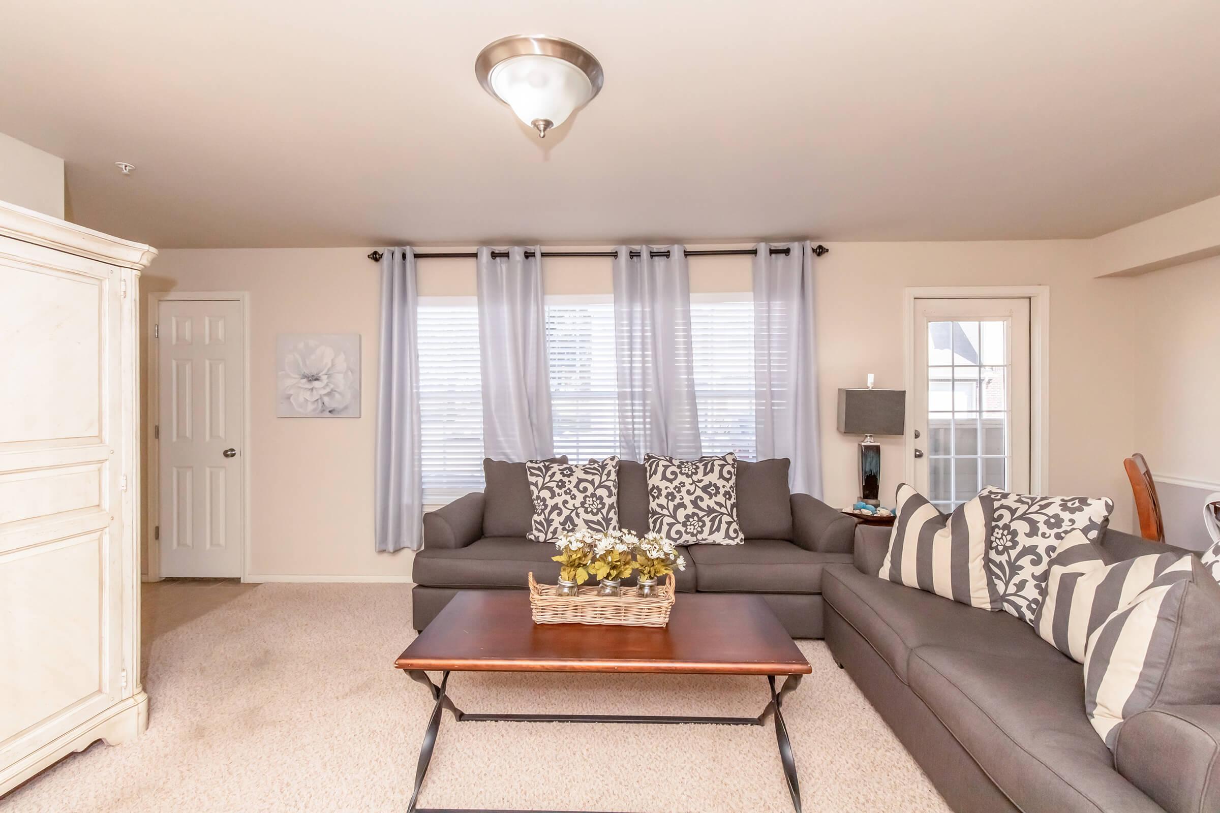 A cozy living room featuring a gray sofa with decorative pillows, a wooden coffee table with a basket of flowers, and light-colored walls. Large windows with sheer curtains allow natural light to enter. A decorative lamp and a white wardrobe add charm to the space, creating a welcoming atmosphere.