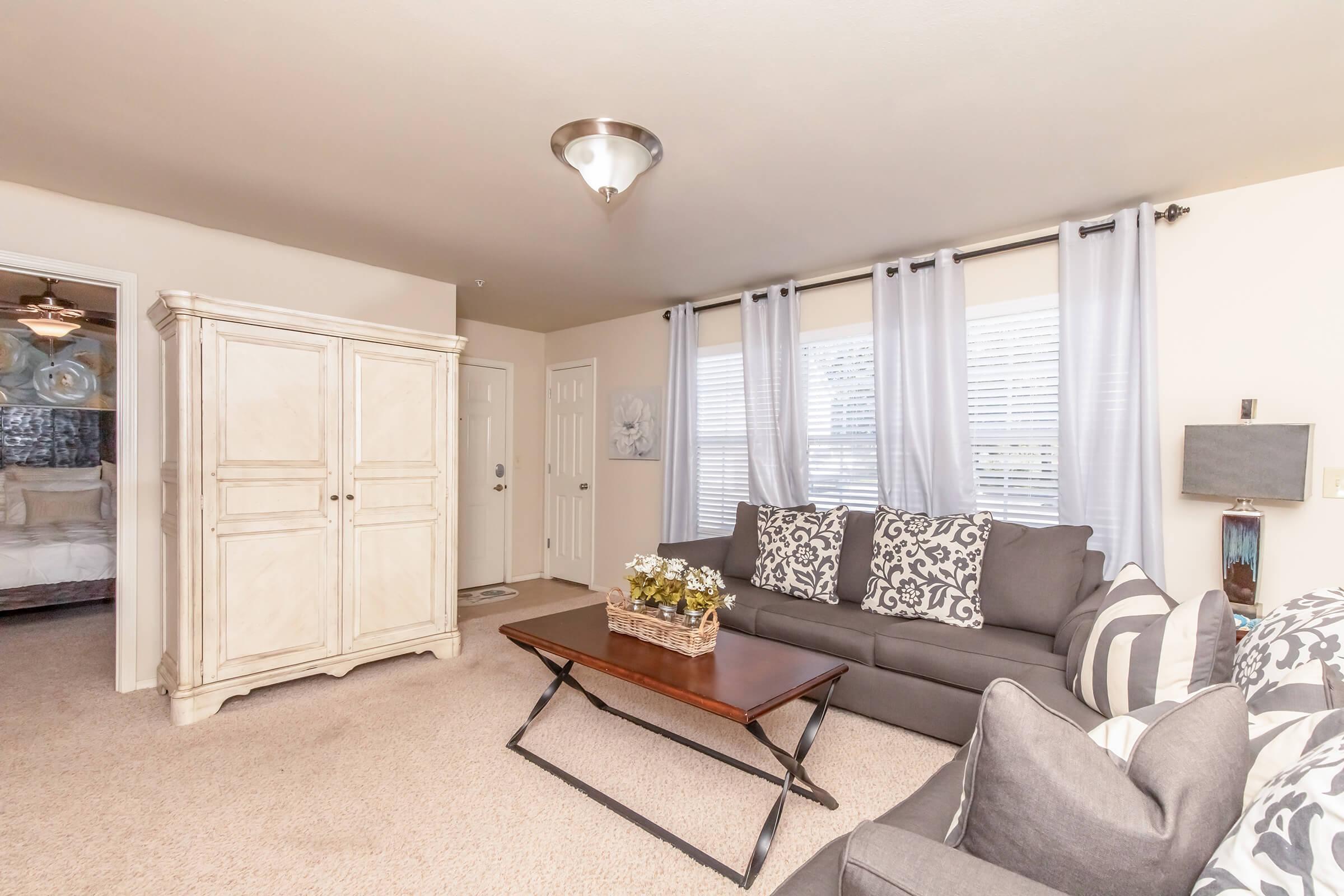 A cozy living room with a gray sofa adorned with patterned pillows. A wooden coffee table sits in front, topped with a decorative vase of flowers. Sheer curtains frame the windows, allowing natural light to fill the space. An armoire and a door to another room are visible in the background.