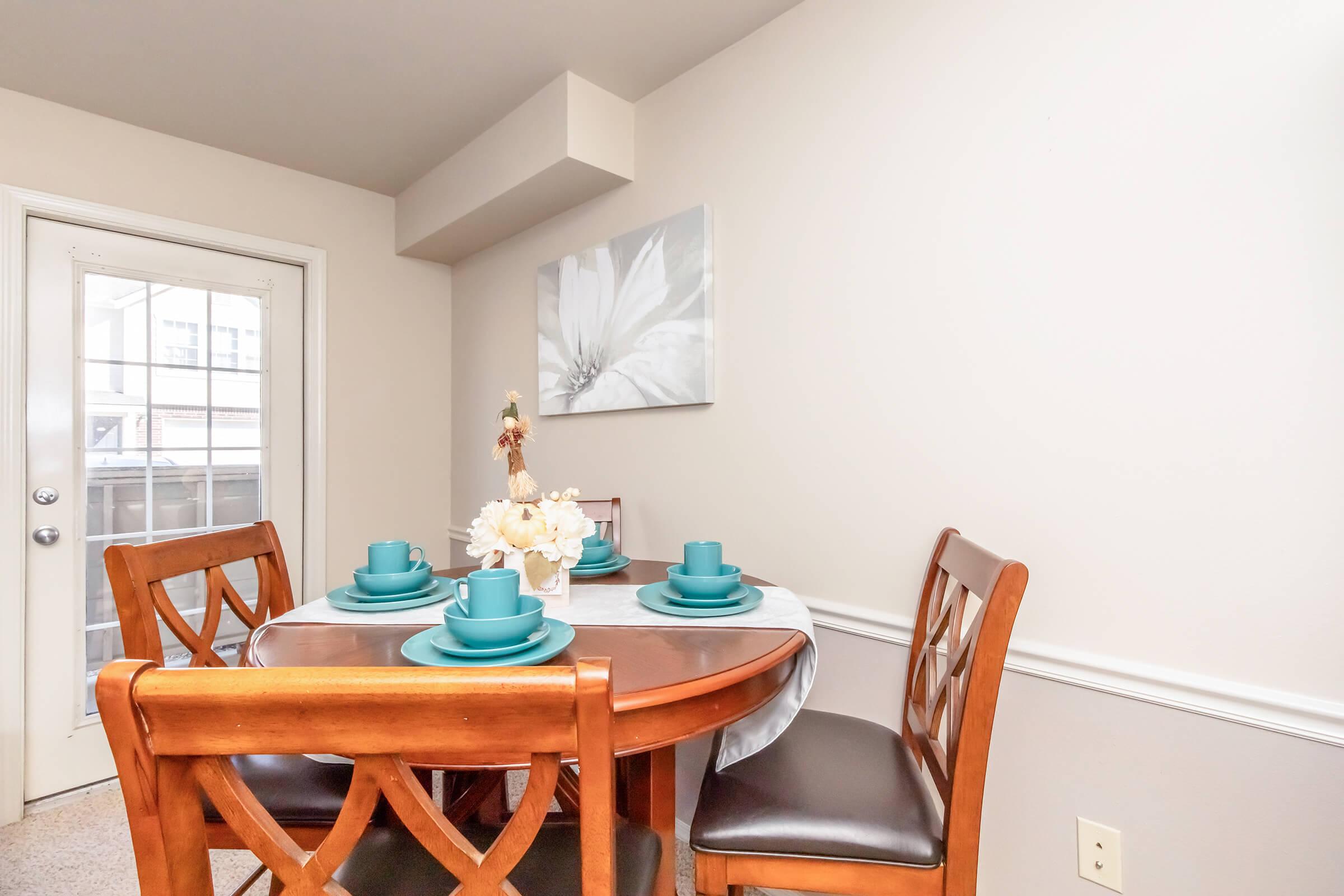 A cozy dining area featuring a round wooden table set for four with turquoise dishes. A small vase with white flowers sits in the center. The walls are light-colored, and a large floral artwork adorns one wall. A doorway leads to an outside area, adding natural light to the space.