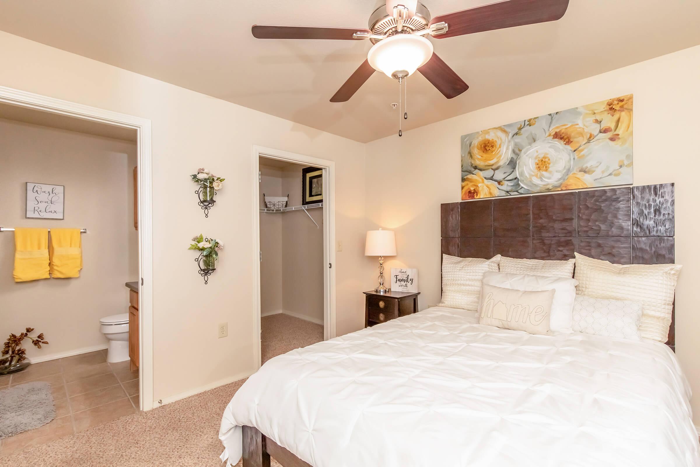 A cozy bedroom featuring a queen-sized bed with decorative pillows, a ceiling fan, and floral artwork above the headboard. The room includes a nightstand with a lamp and has an open doorway leading to a bathroom and a closet. Soft, neutral tones create a warm and inviting atmosphere.