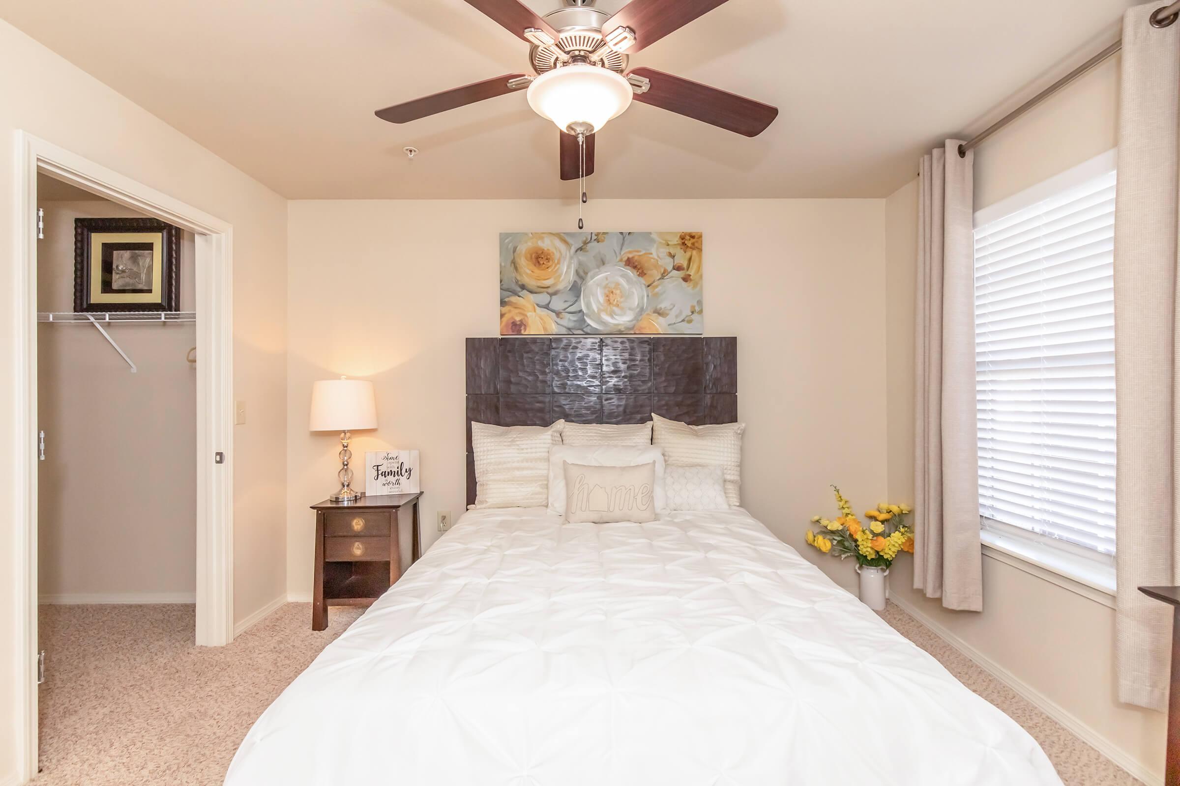 Cozy bedroom featuring a large bed with white bedding and decorative pillows. A wooden headboard complements the light-colored walls. A window with light curtains lets in natural light, while a small nightstand with a lamp and a framed picture adds charm. Fresh yellow flowers enhance the inviting atmosphere.