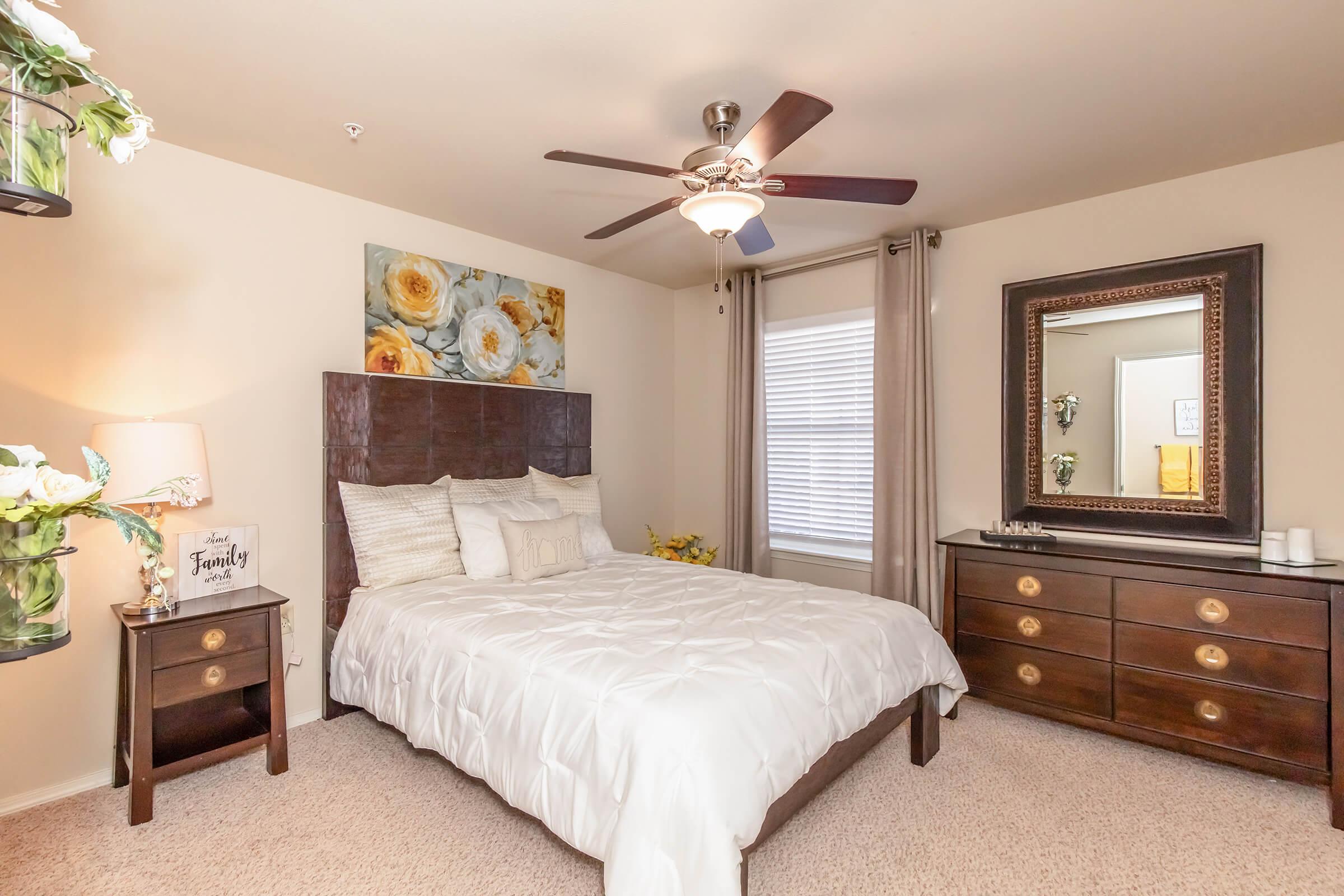 A cozy bedroom featuring a queen-sized bed with a white quilt, decorative pillows, and a wood headboard. There’s a large mirror above a dark wood dresser, and a ceiling fan with light. Soft curtains frame a window, and a lamp with a floral arrangement adds warmth to the space. The walls are painted in neutral tones.