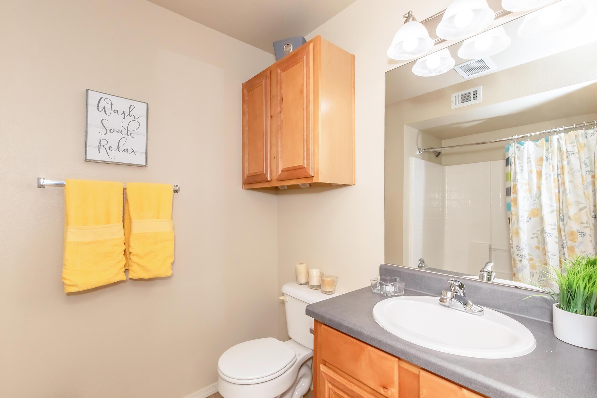 A bright bathroom featuring light beige walls, a wooden vanity with a gray countertop, a white toilet, and a shower curtain with floral patterns. Two yellow towels hang on a rack, and there are decorative candles on the counter. A wall sign reads "Wash, Soak, Relax." Natural light illuminates the space.
