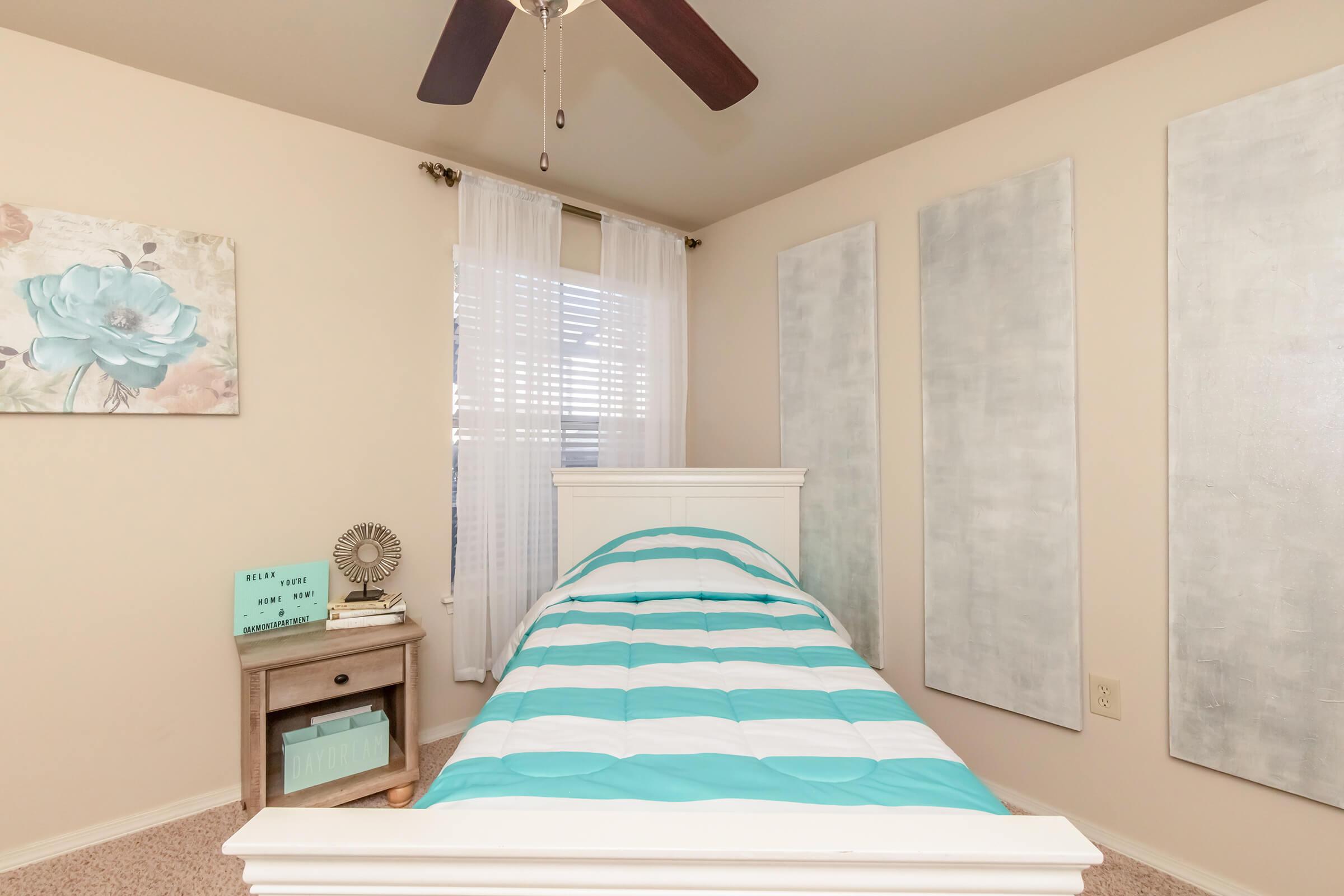 A cozy bedroom featuring a single bed with a striped turquoise and white comforter, pale walls, and two abstract wall panels. A nightstand with a decorative fan and a small turquoise sign is beside the bed. Sheer white curtains allow natural light to filter in, creating a warm and inviting atmosphere.