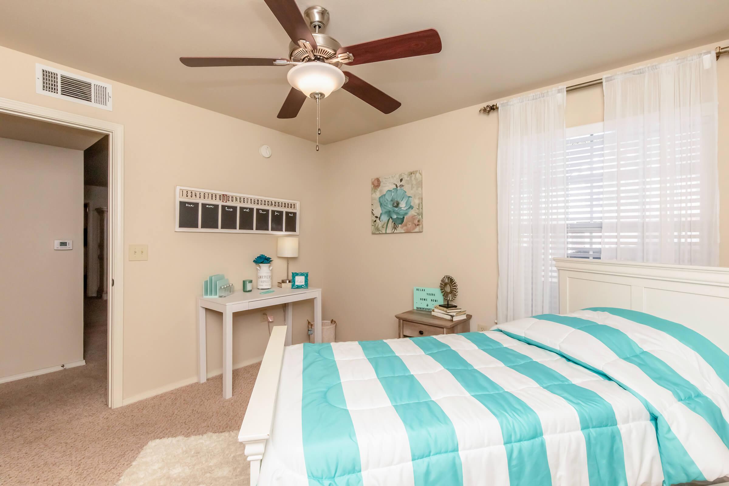 Brightly lit bedroom featuring a white bed with a turquoise and white striped comforter. A small desk with decorative items is placed next to the bed, along with a nightstand. The walls are a soft beige, and there's a ceiling fan overhead. Sheer curtains adorn the window, allowing natural light to filter in.