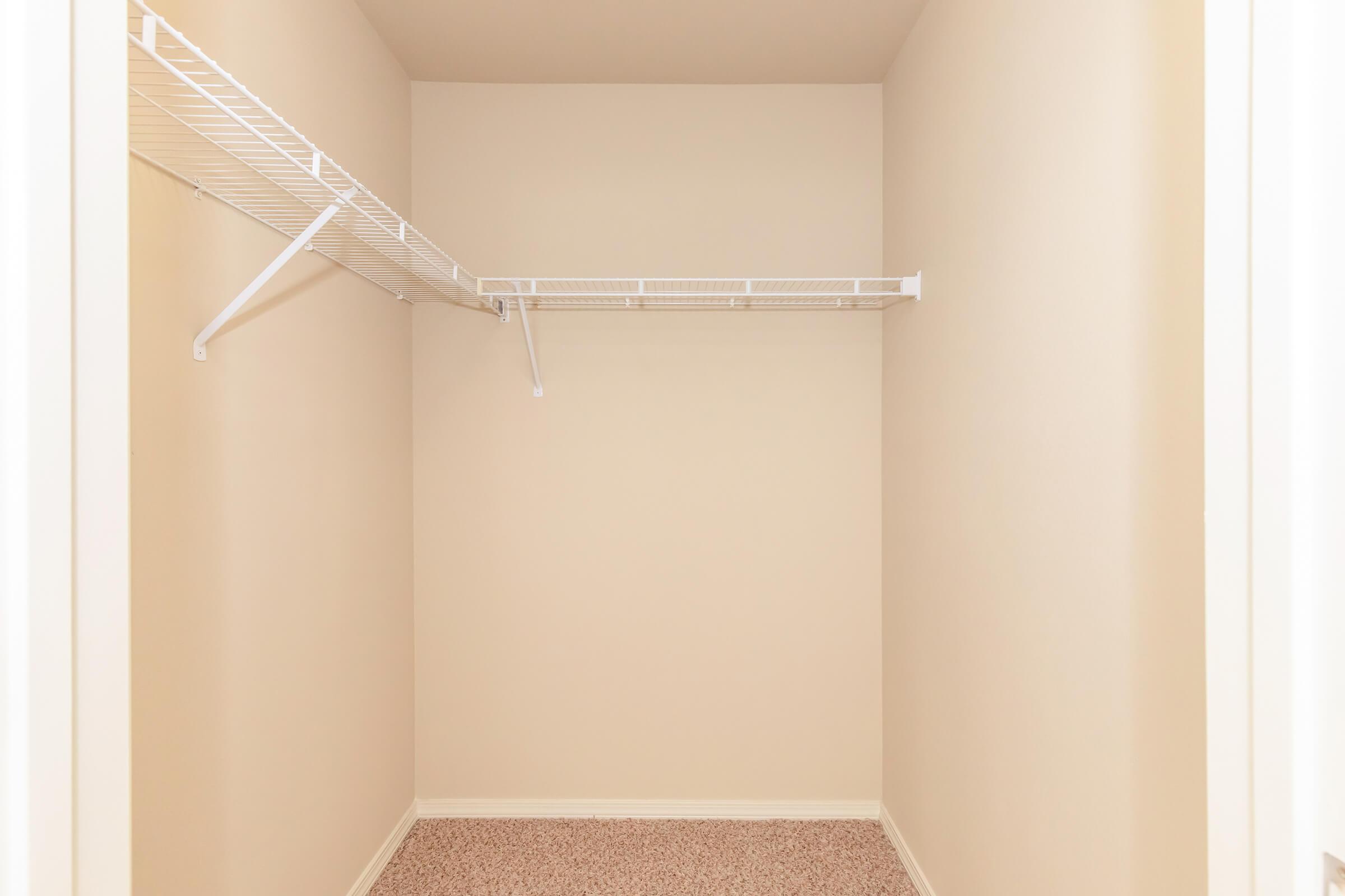 Empty closet with beige walls and carpeted floor. The closet features a white wire shelf along the back wall, with an additional hanging rod extending from one side. The overall space is minimal and uncluttered, providing a simple and clean storage area.