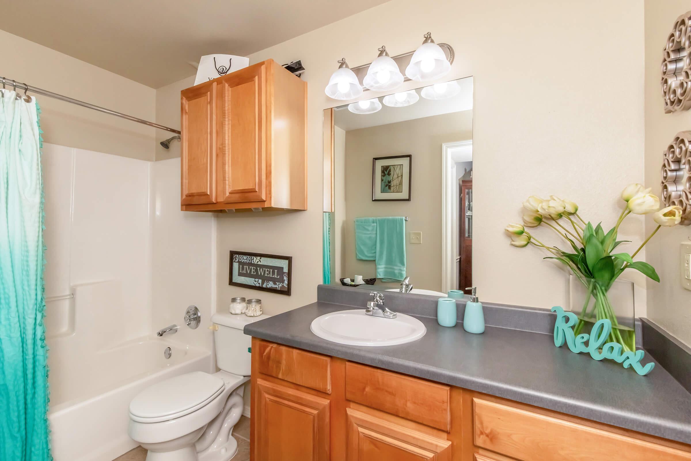 A modern bathroom featuring a light wood vanity with a gray countertop, a white sink, and a bath/shower combo with a turquoise curtain. Decorative elements include a mirror, a vase with flowers, and a sign that reads "Relax." The overall color scheme is soft and inviting.