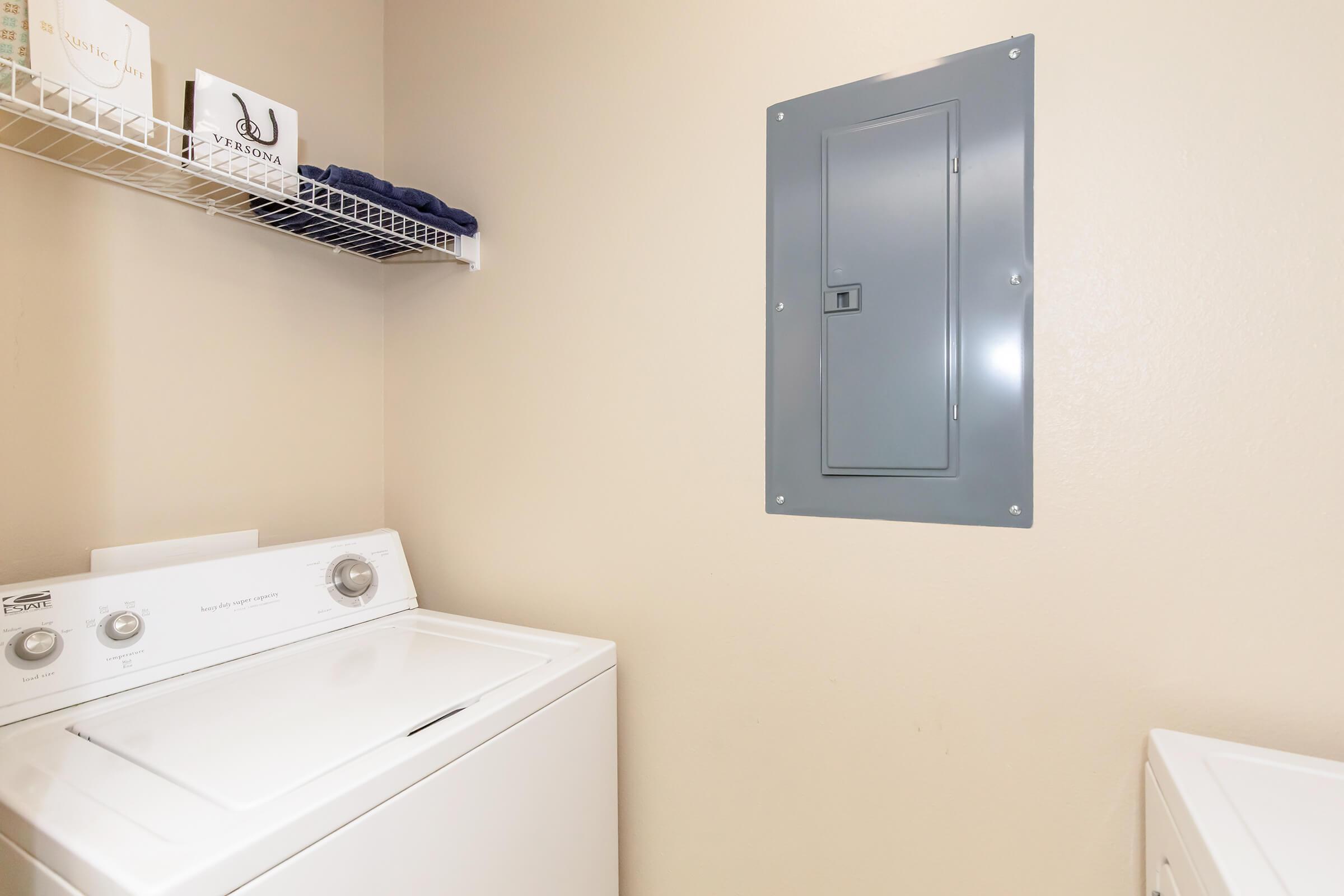 A laundry room featuring a white washing machine, a shelf with folded towels, and an electrical panel on the beige wall. The floor is light-colored, creating a tidy and functional space.