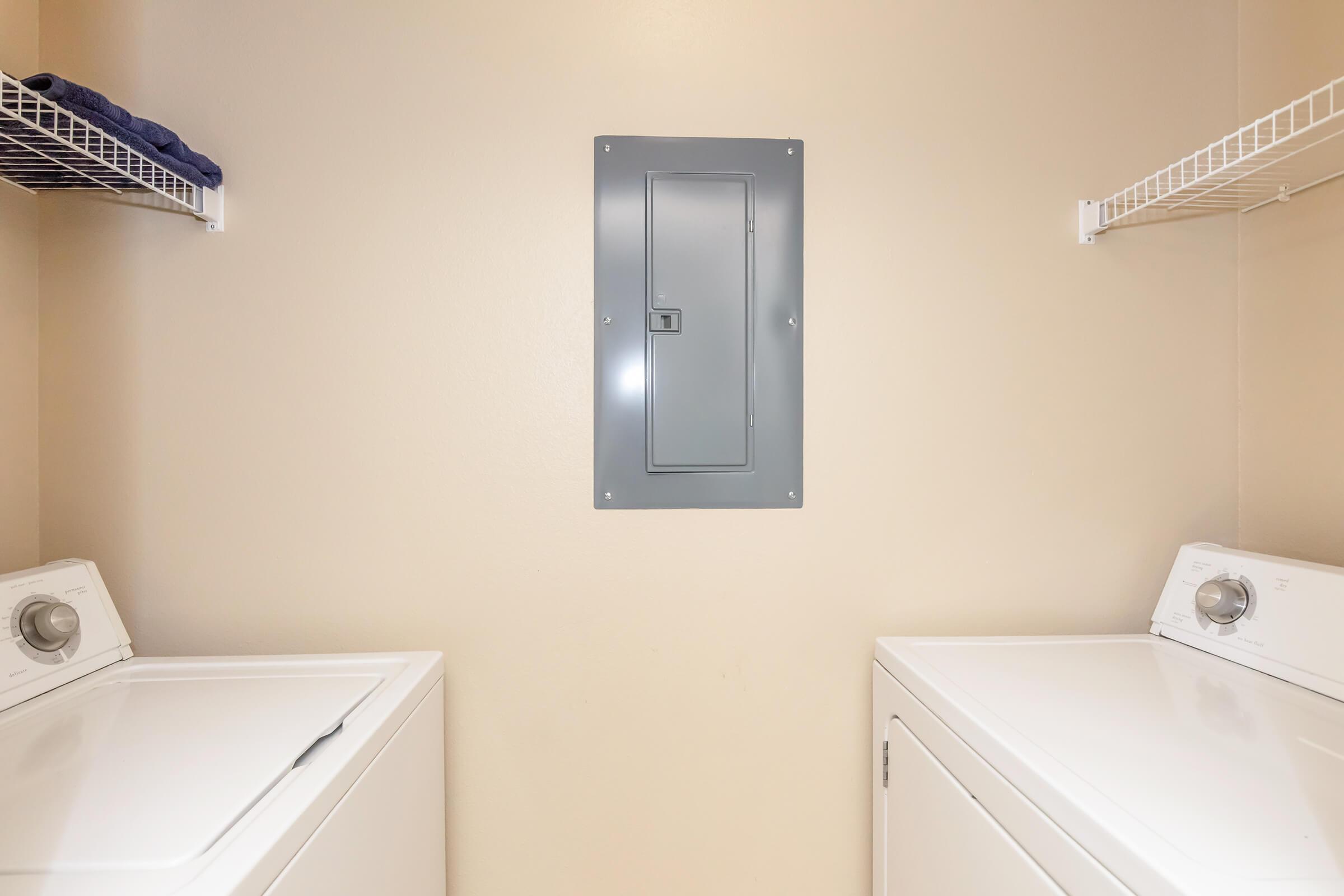A laundry room featuring two white washing machines side by side, with a beige wall in the background. Above each machine, there are wire shelves stocked with folded towels. A gray electrical panel is mounted on the wall between the machines. The setting is clean and organized, with neutral colors.