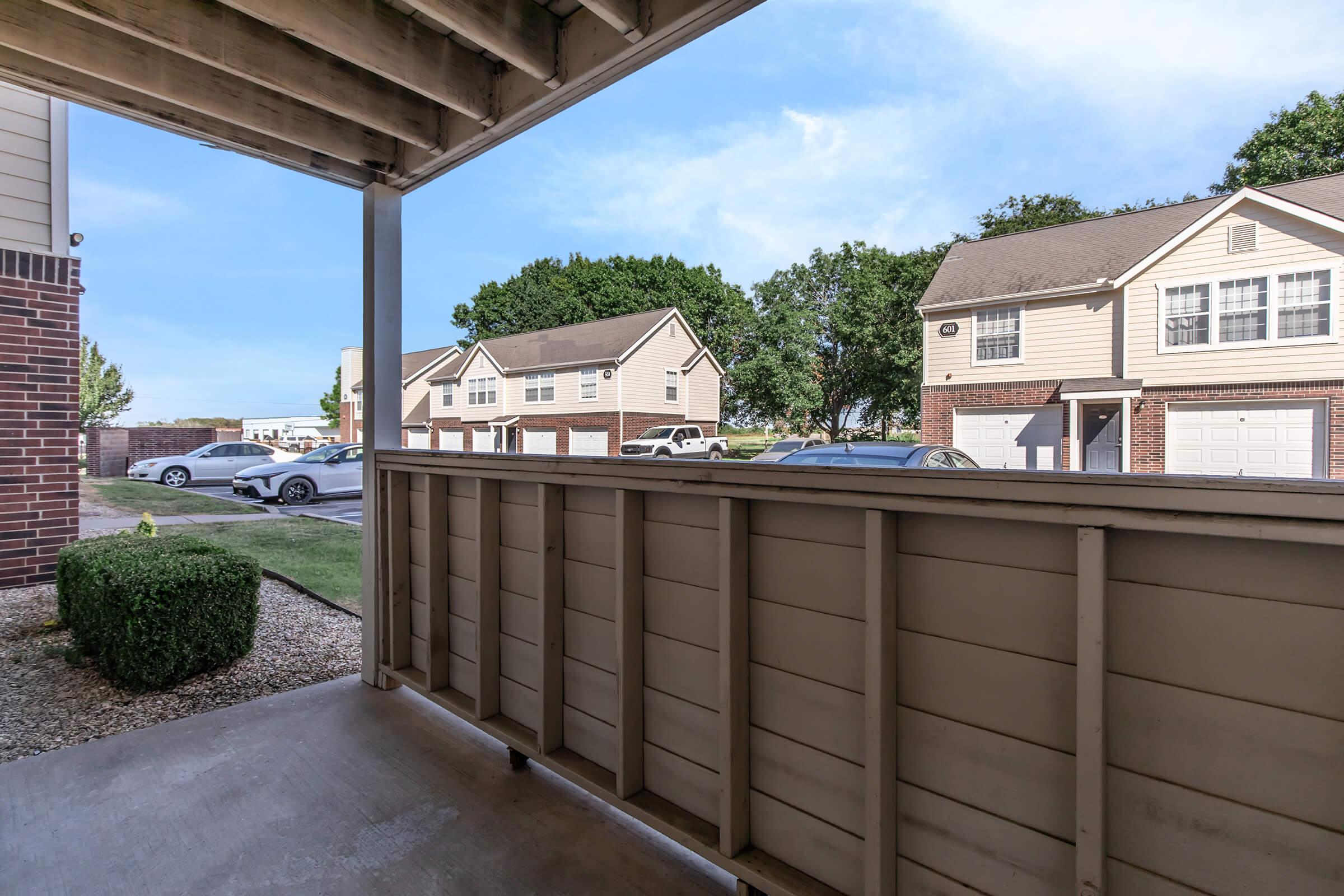 View from a covered porch looking out towards a residential area. Features a wooden railing, a patch of grass, and several parked cars. In the background, rows of townhouses are visible under a clear blue sky.