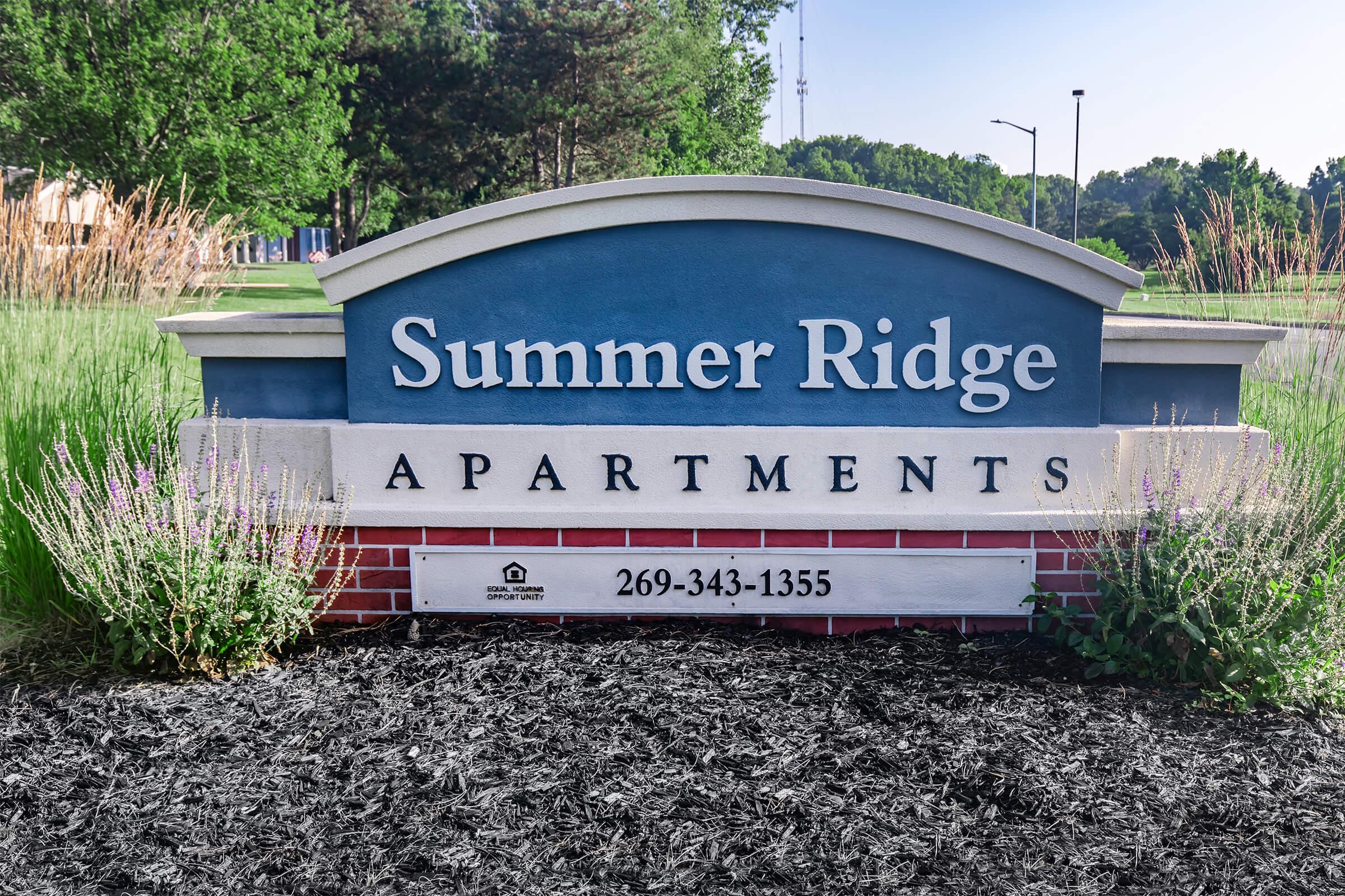 Sign for Summer Ridge Apartments, featuring the name prominently displayed. Below the name, a phone number (269-343-1355) is visible. The sign is set against a backdrop of greenery, with ornamental plants and a dark mulch base.