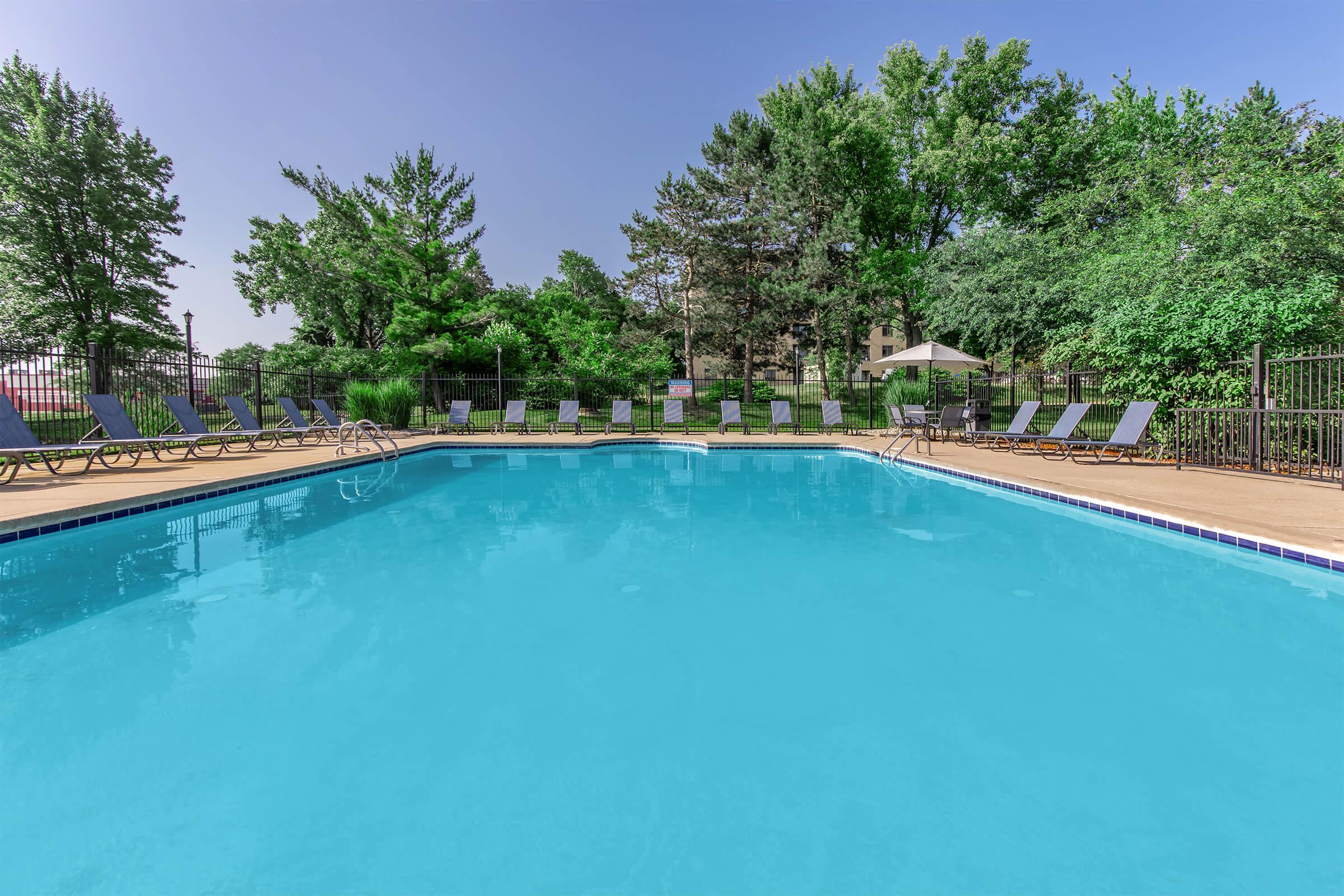 A clear swimming pool surrounded by lounge chairs, set in a lush green garden. Tall trees and shrubs provide a natural backdrop, and the sky is bright blue with minimal clouds. Sunlight reflects off the water, creating a tranquil and inviting atmosphere for relaxation.
