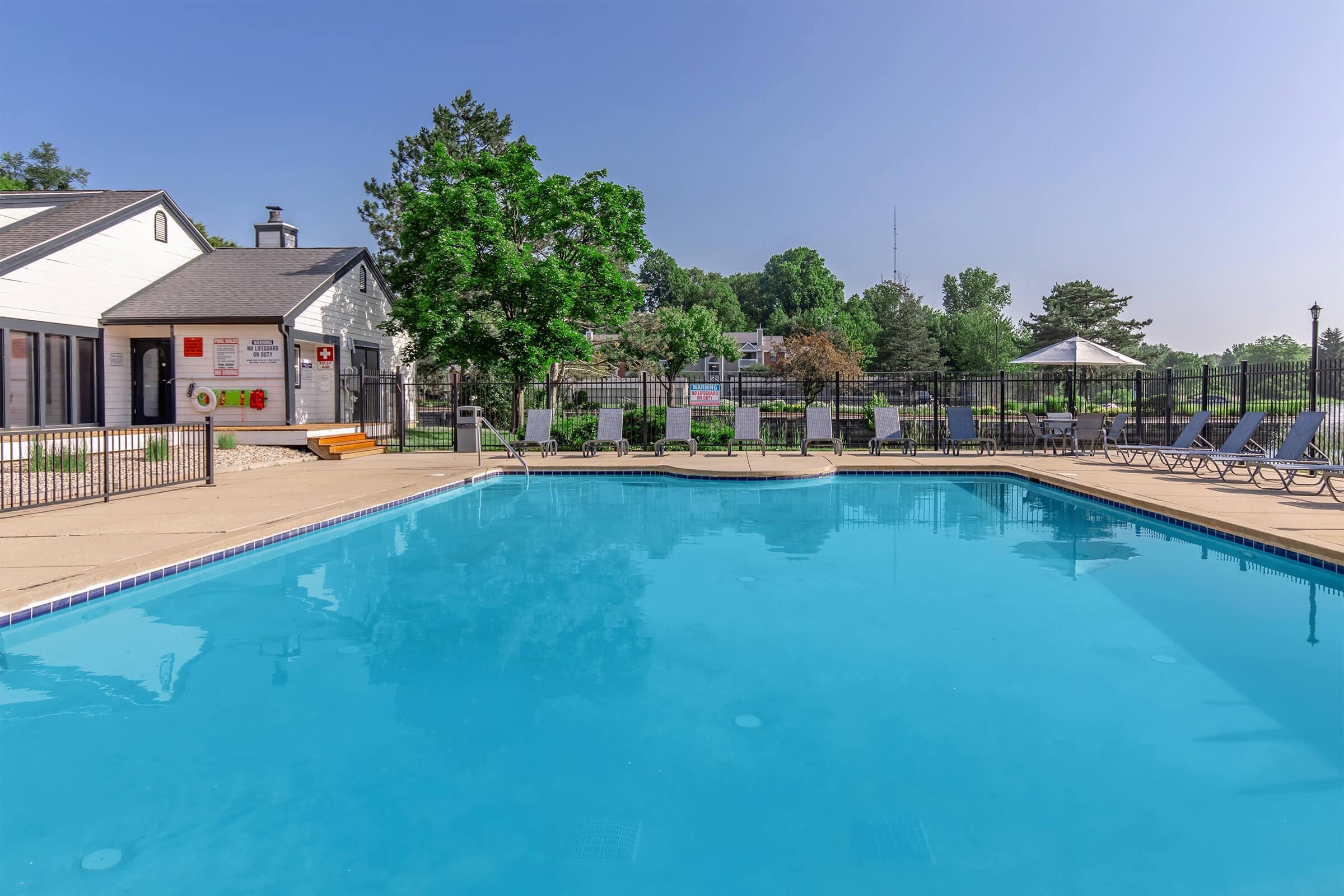 A clear blue swimming pool surrounded by a patio area with lounge chairs. In the background, there are trees and a fence, along with a small building that has signs. The sky is bright and clear, indicating a sunny day. The atmosphere appears calm and inviting.