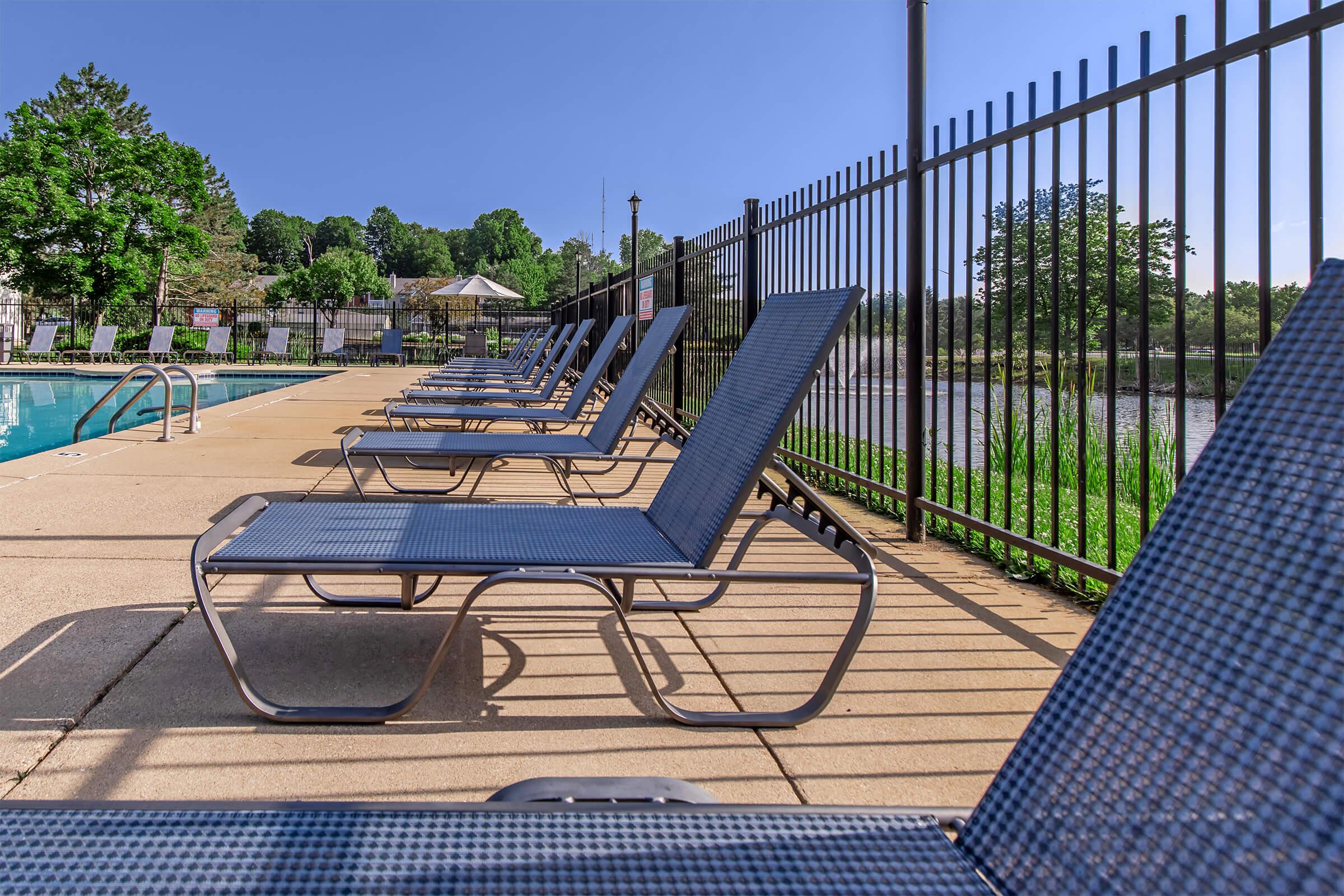 A row of blue lounge chairs lined up beside a swimming pool, with a clear sky in the background. The area is enclosed by a black fence, and greenery is visible around the pool, creating a tranquil outdoor setting.
