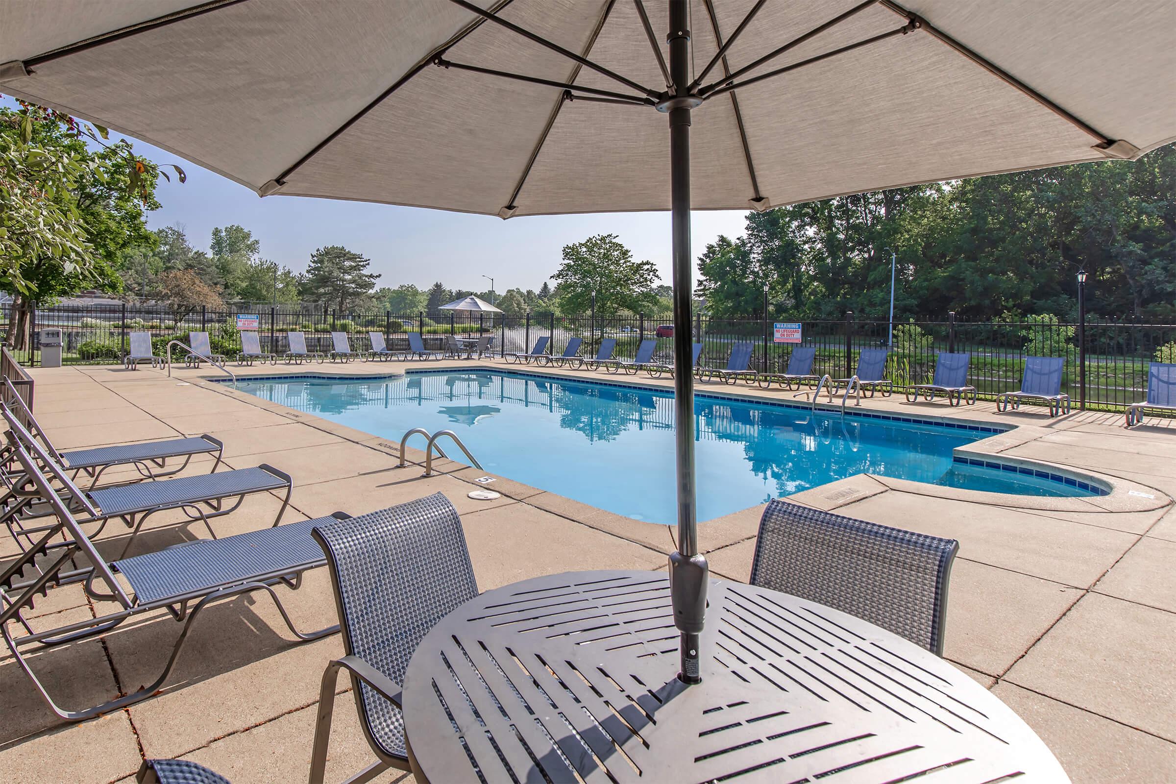 A view of a pool area featuring a large swimming pool surrounded by lounge chairs and a shaded table with an umbrella. Lush greenery and trees are visible in the background, providing a relaxing atmosphere. The pool has a diving board on one side, and there are signs around the area.
