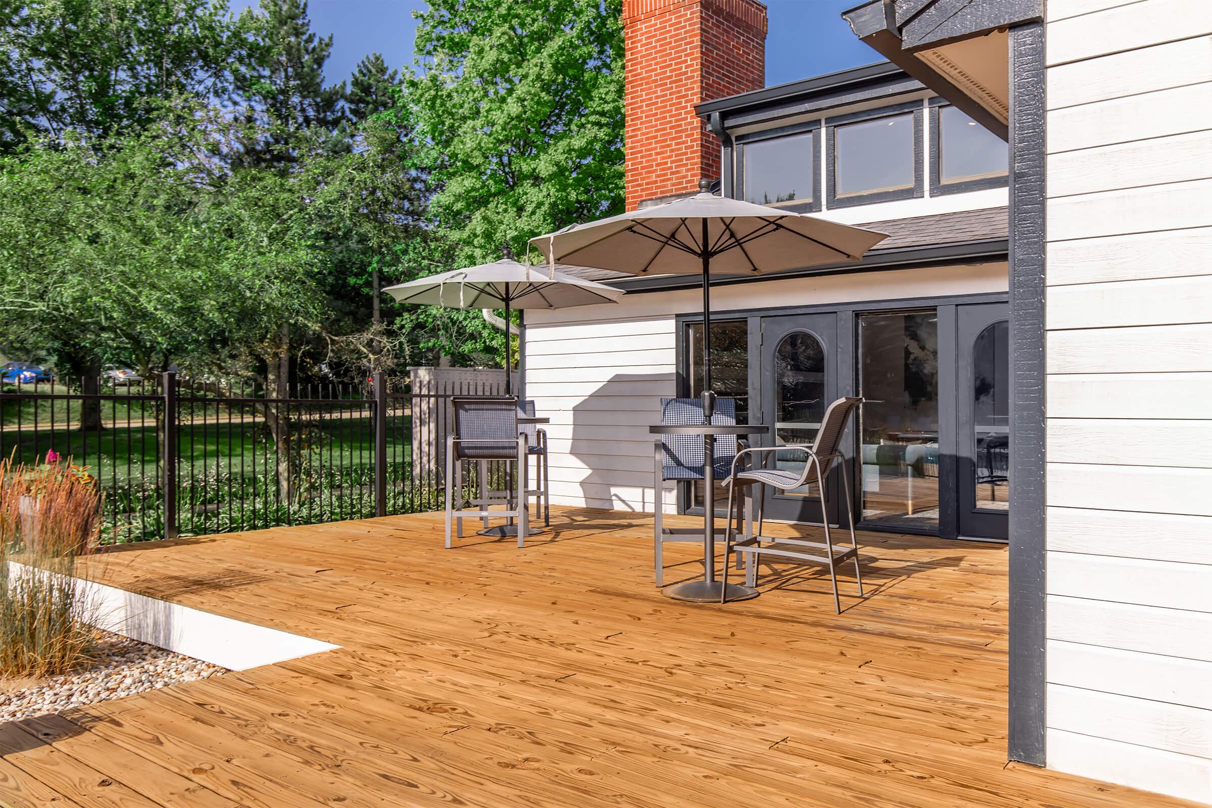 A sunny outdoor deck featuring wooden flooring, two gray patio chairs, a small table, and two large umbrellas. In the background, a modern house with large windows and a chimney is visible, surrounded by lush green trees and a fenced yard.