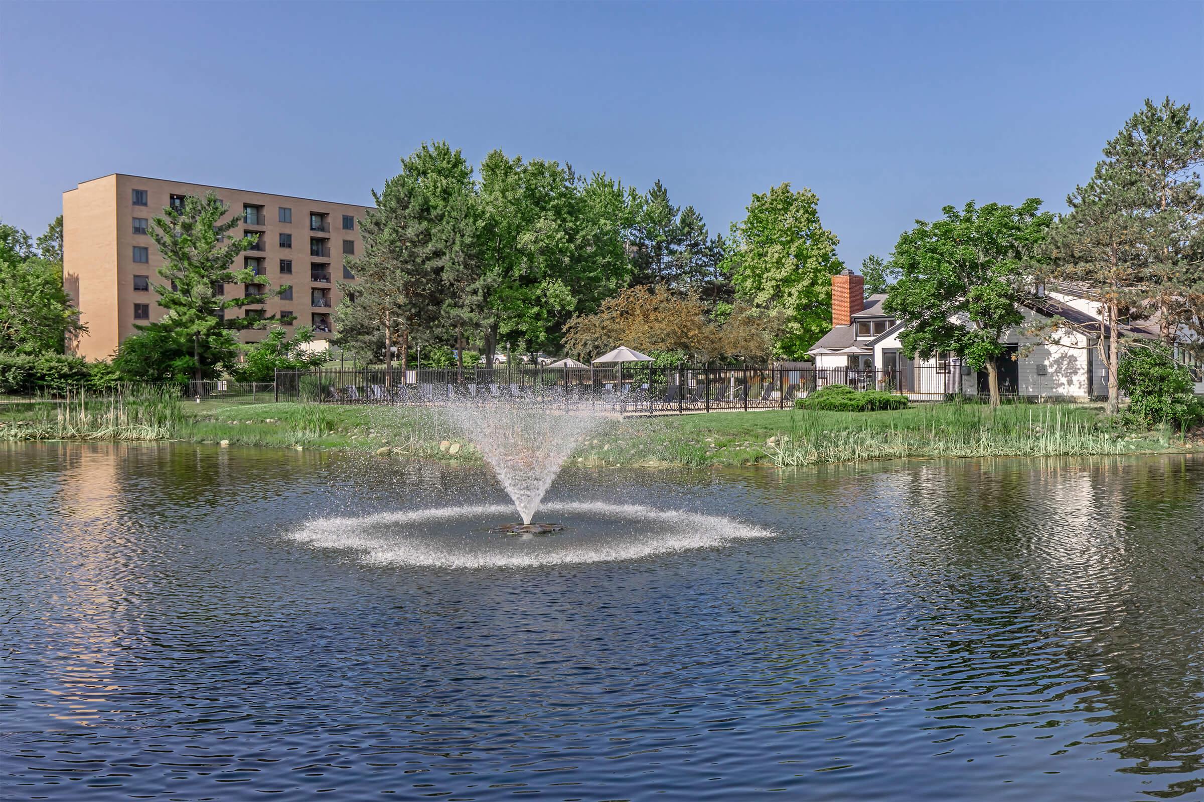 A serene pond with a water fountain in the center, surrounded by lush green trees and grass. In the background, there is a modern building and a small white house, adding to the peaceful atmosphere of the scene. Clear blue sky reflects in the water, enhancing the tranquil setting.