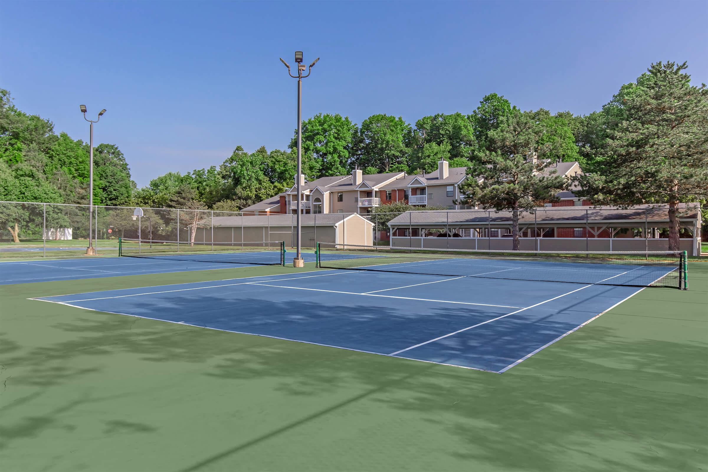A view of several tennis courts with blue surfaces and white lines, set against a backdrop of green trees and residential buildings. The sky is clear and blue, with a few light clouds. Bright lights are positioned above the courts for nighttime use.