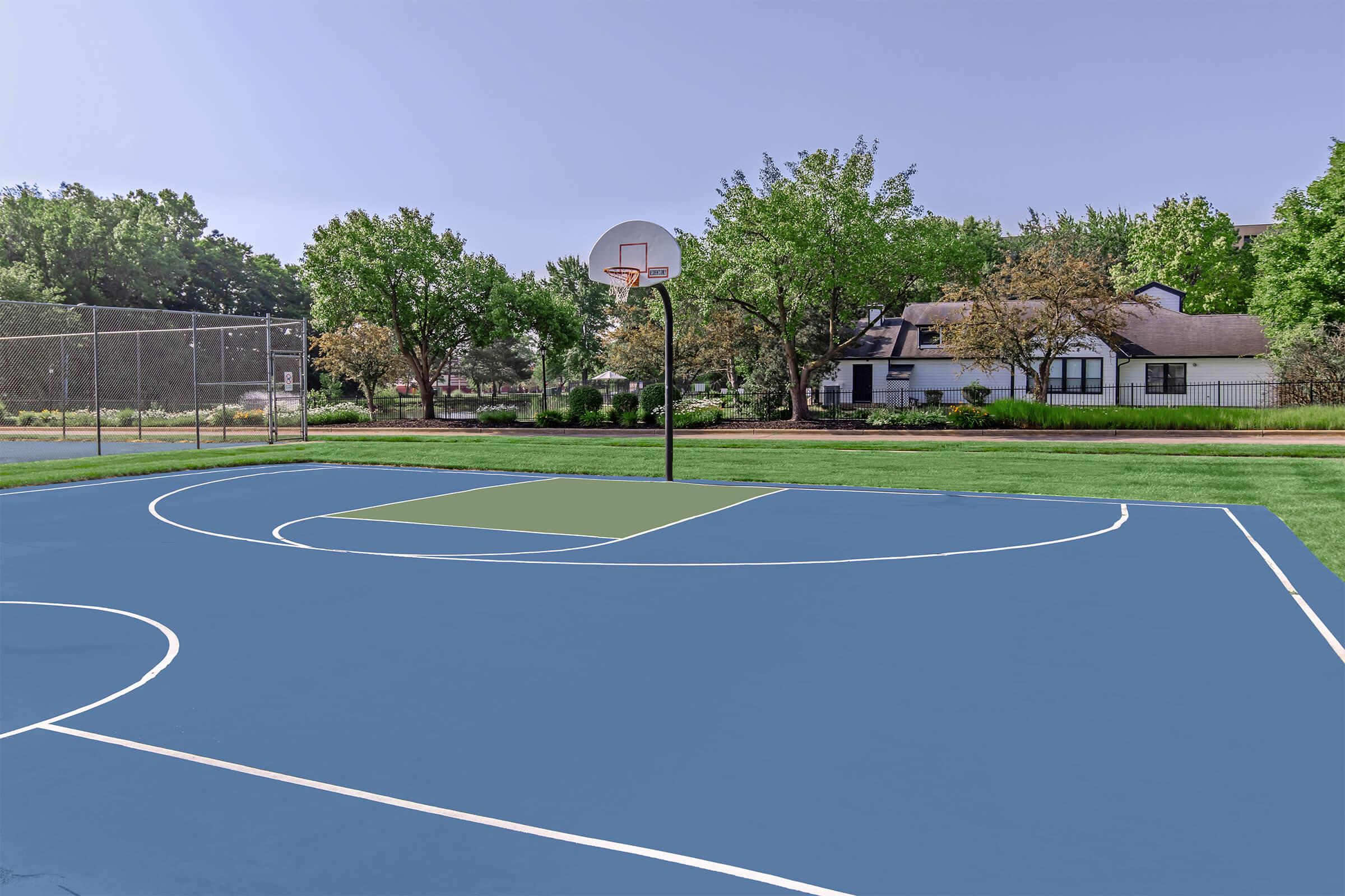 A well-maintained outdoor basketball court with a blue surface and marked lines, surrounded by lush green trees and a residential area in the background. A basketball hoop is visible at one end of the court, with a chain-link fence enclosing another area nearby.