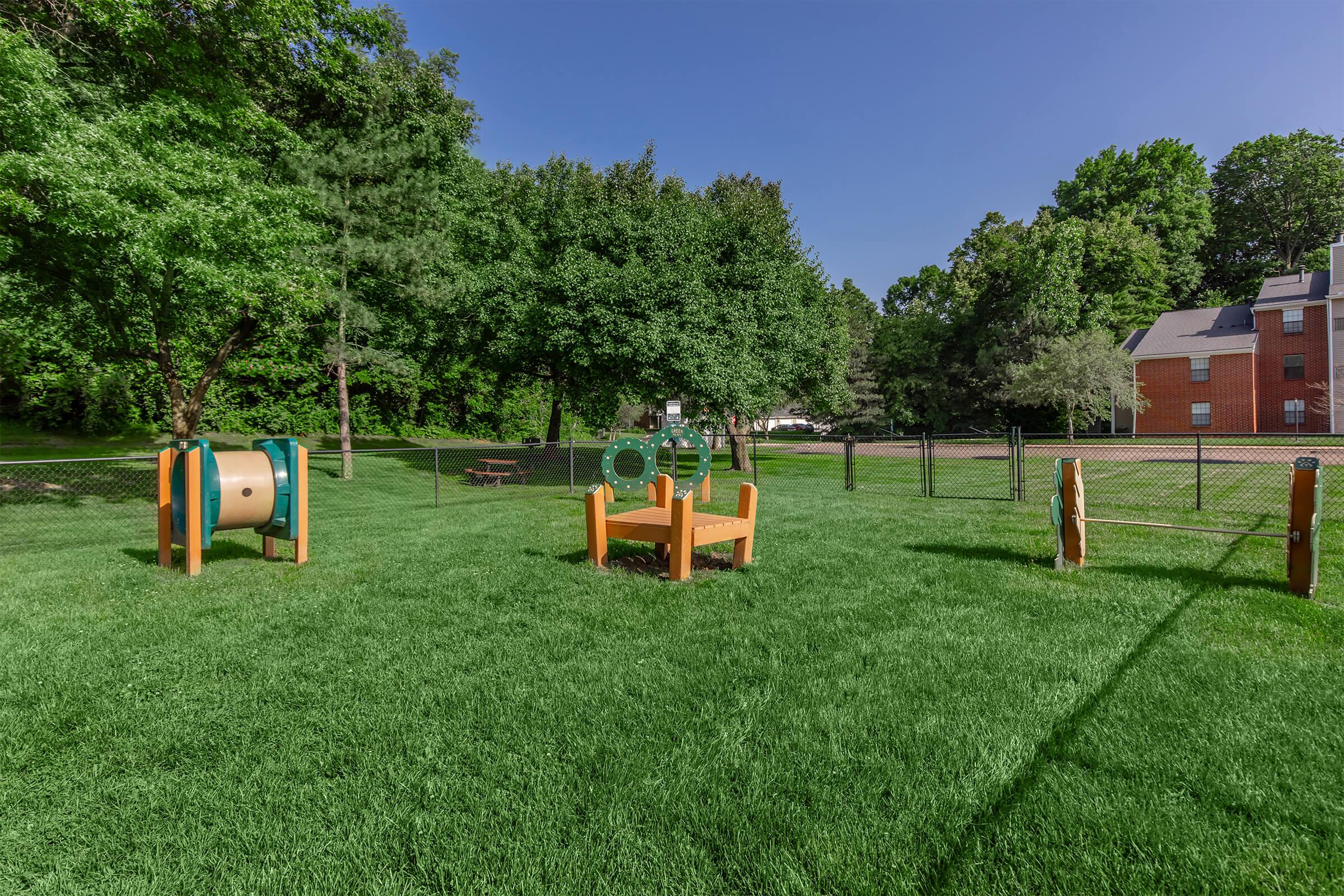 A grassy playground area with colorful outdoor equipment, including a circular structure and a drum, surrounded by trees and a fence. A building can be seen in the background, and the sky is clear and blue. The space appears inviting and suitable for children to play.