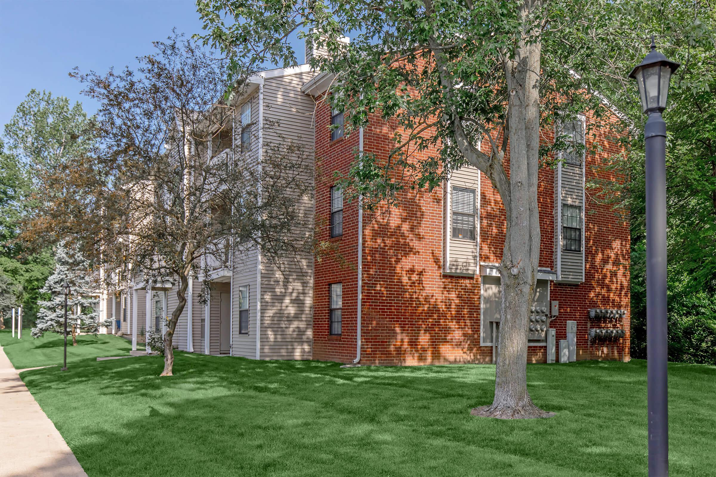 A residential building with a mix of brick and wood siding, surrounded by a green lawn and trees. The setting is sunny, and there is a pathway leading alongside the building. A streetlamp is visible on the right side of the image.