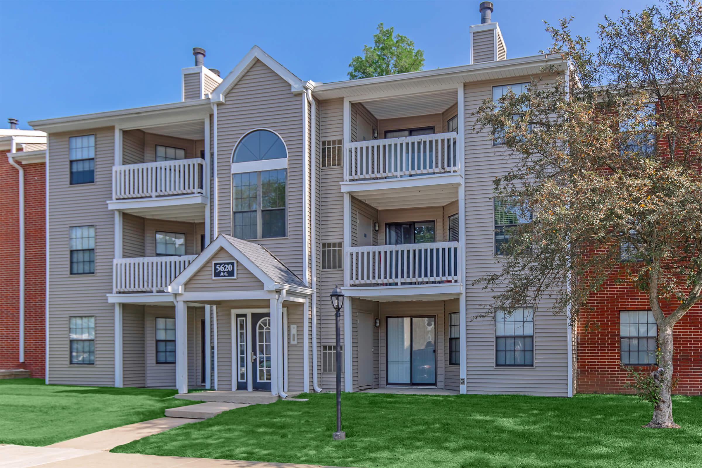 A multi-story apartment building with a light beige exterior featuring balconies on the second floor. The entrance is centered, with large windows flanking the door. There is a green lawn in front, and a lamp post is visible on the walkway leading to the entrance. The sky is clear and blue.