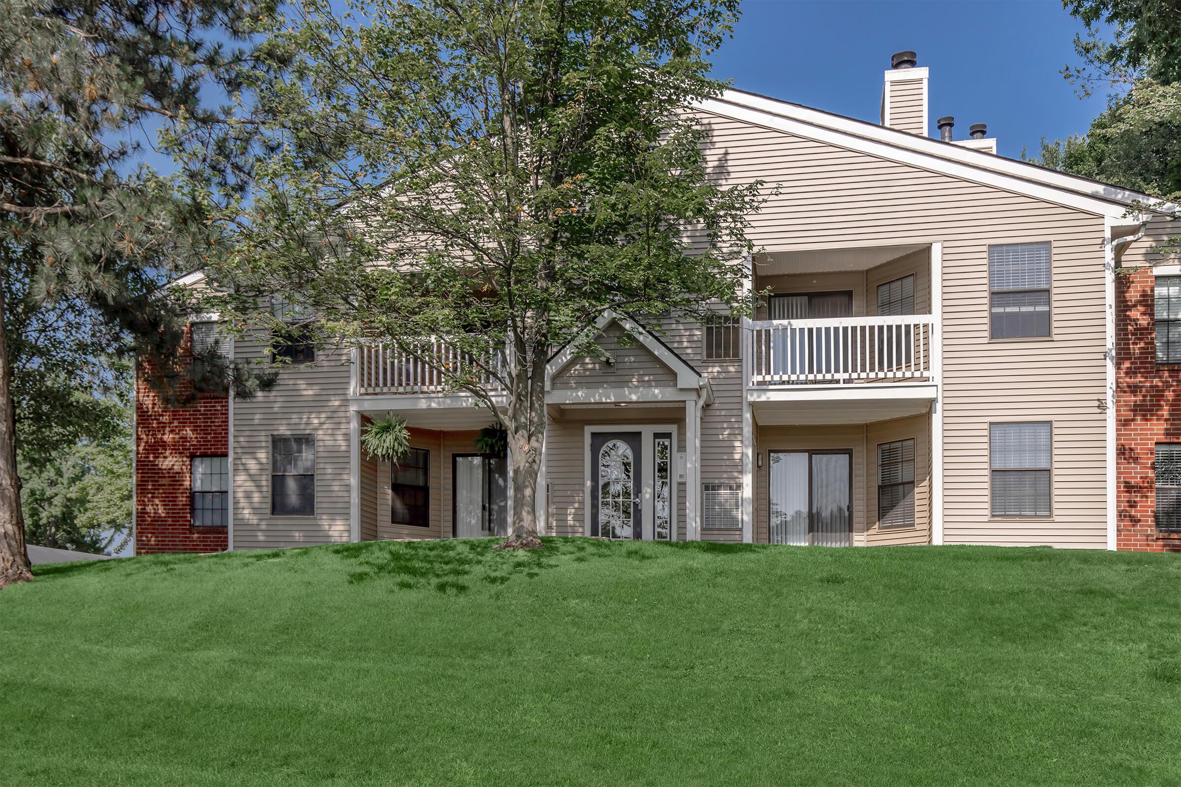 A two-story apartment building with a light-colored exterior and balconies. The building is surrounded by well-maintained green grass and trees, under a clear blue sky. The layout features multiple windows and a front entrance with decorative elements.