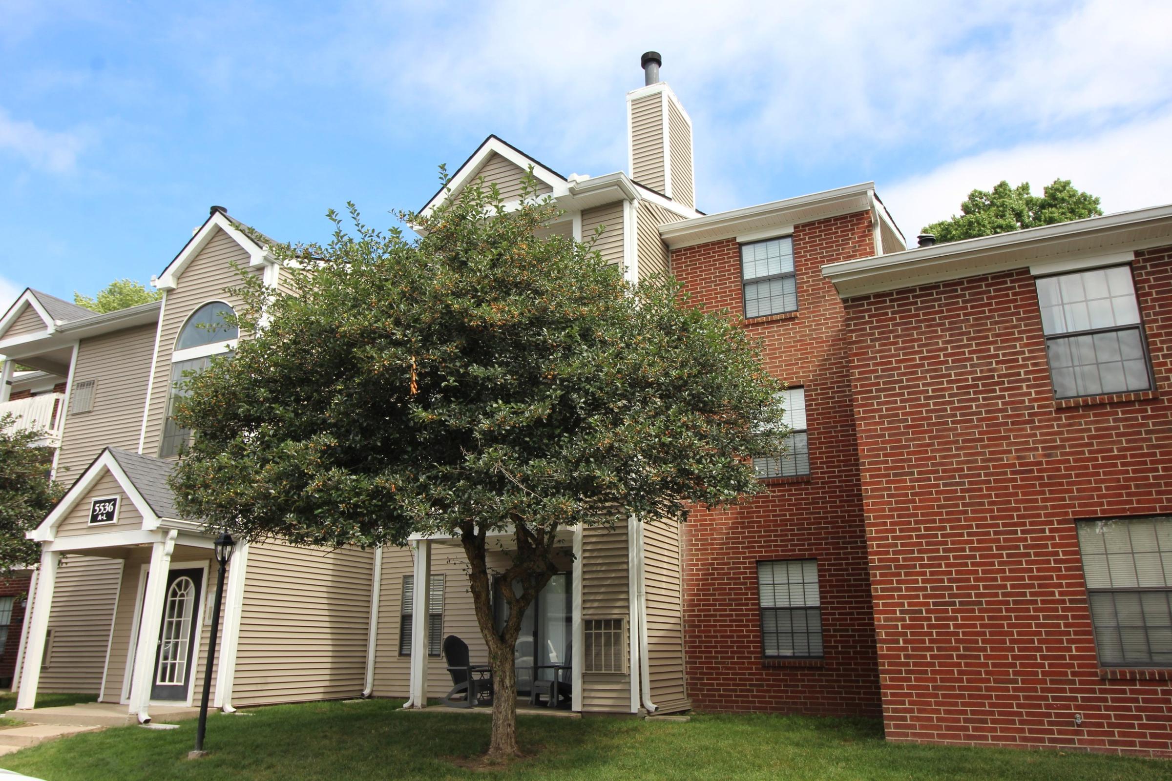 A residential building with a mix of beige siding and red brick, featuring multiple windows and a tree in front. The structure includes a chimney and a small lamp post beside a green lawn. The sky is partly cloudy, providing a bright ambiance.